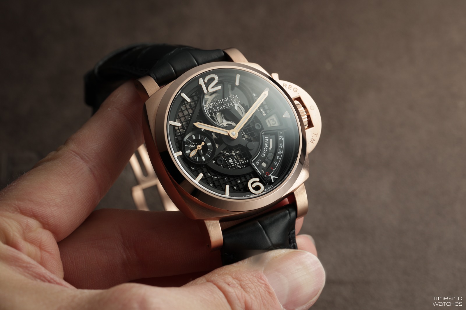 PAM01631 shown held in hand, three-quarter dial view in Goldtech with the crown-protector lever lock visible and REG/T.M. engraved on the crown guard, skeletonised dial showing the 31 GIORNI retrograde scale at 3 o'clock