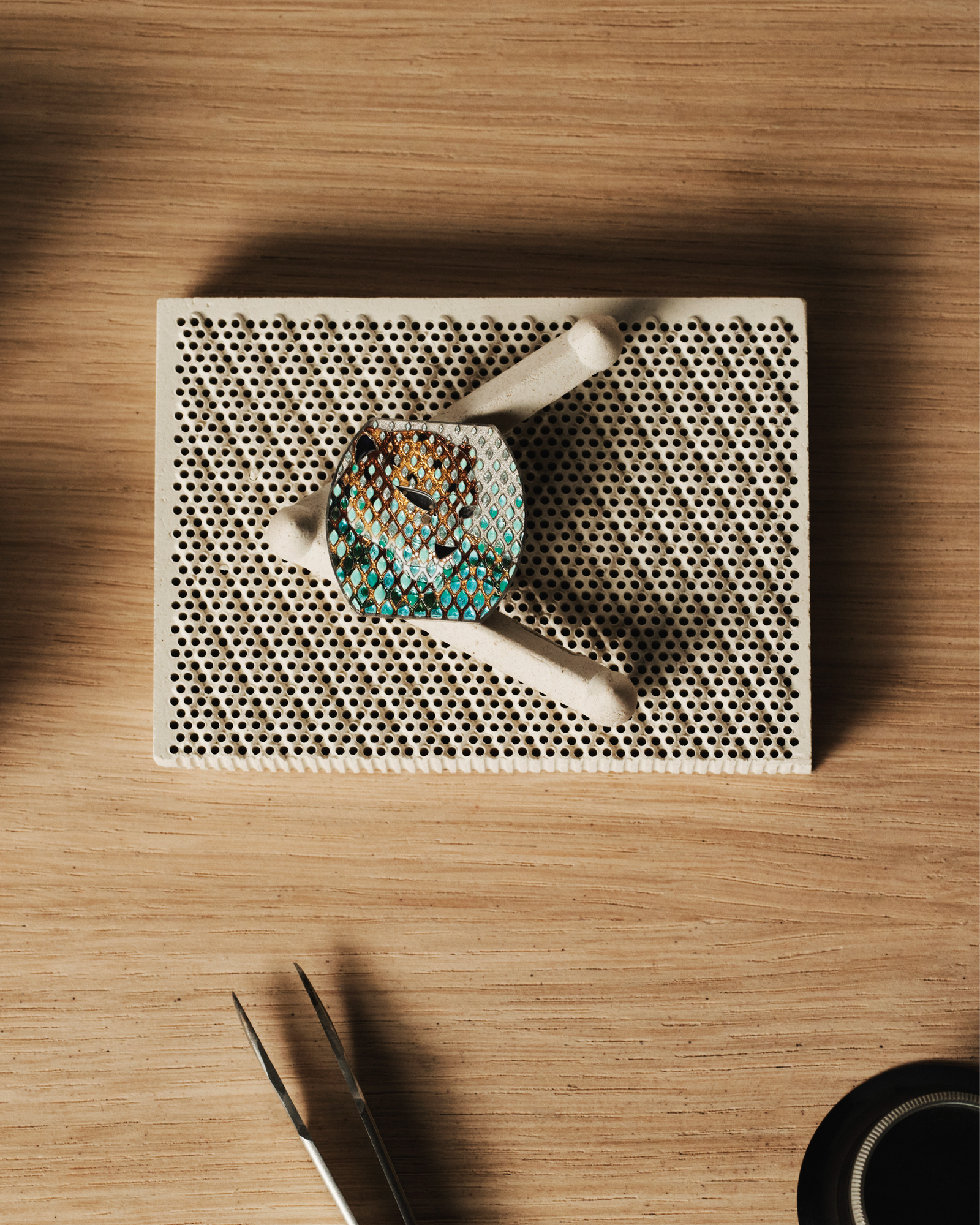 Making-of shot of the Panthère Métiers d'Art Tortue on a pierced brass workbench, with the blue-and-green champlevé-enamelled case and tweezers beside