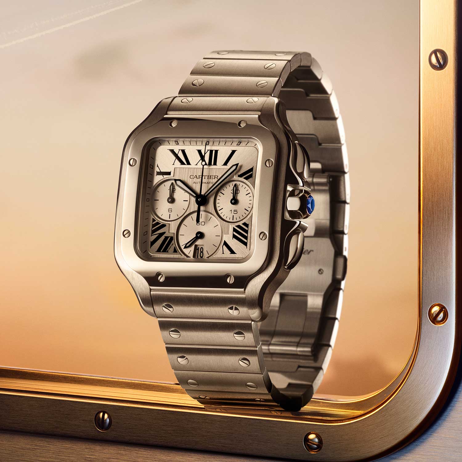 Steel Cartier Santos Chronograph XL 2026 with silver sunburst dial, rhodium-bordered tricompax layout, exposed bezel screws, blue sapphire crown cabochon and steel bracelet, photographed against a brass frame