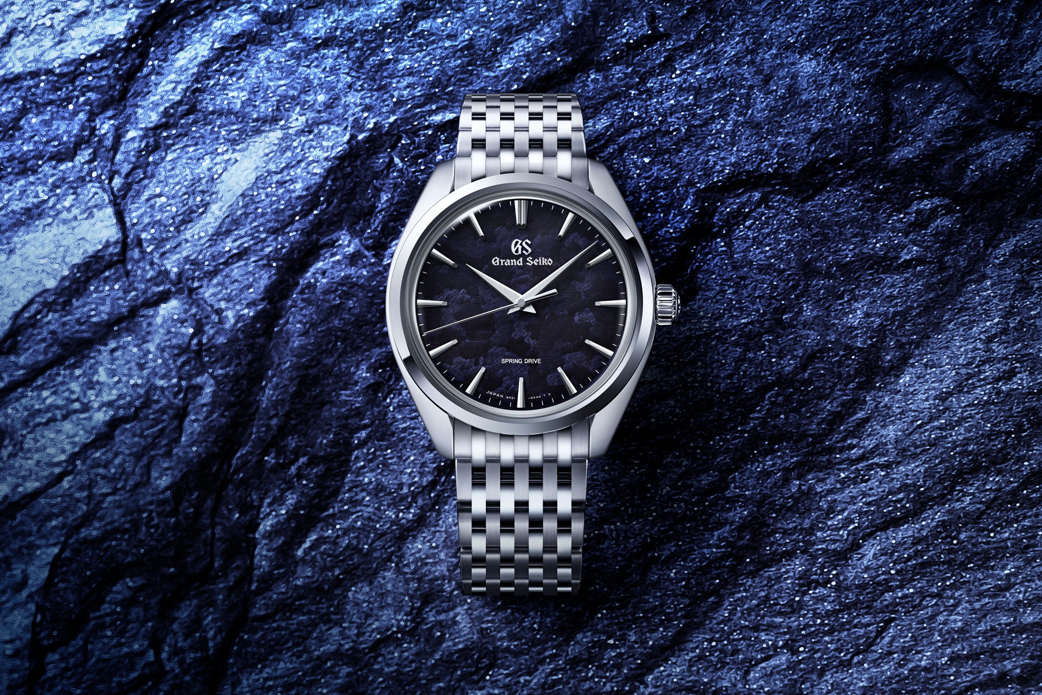 Grand Seiko SBGY043 Iwao Blue in stainless steel, 38.5mm Elegance case on a multi-link steel bracelet, textured indigo dial with applied faceted hour markers, silver hands, Grand Seiko script signature and Spring Drive text, photographed against a textured blue rock surface