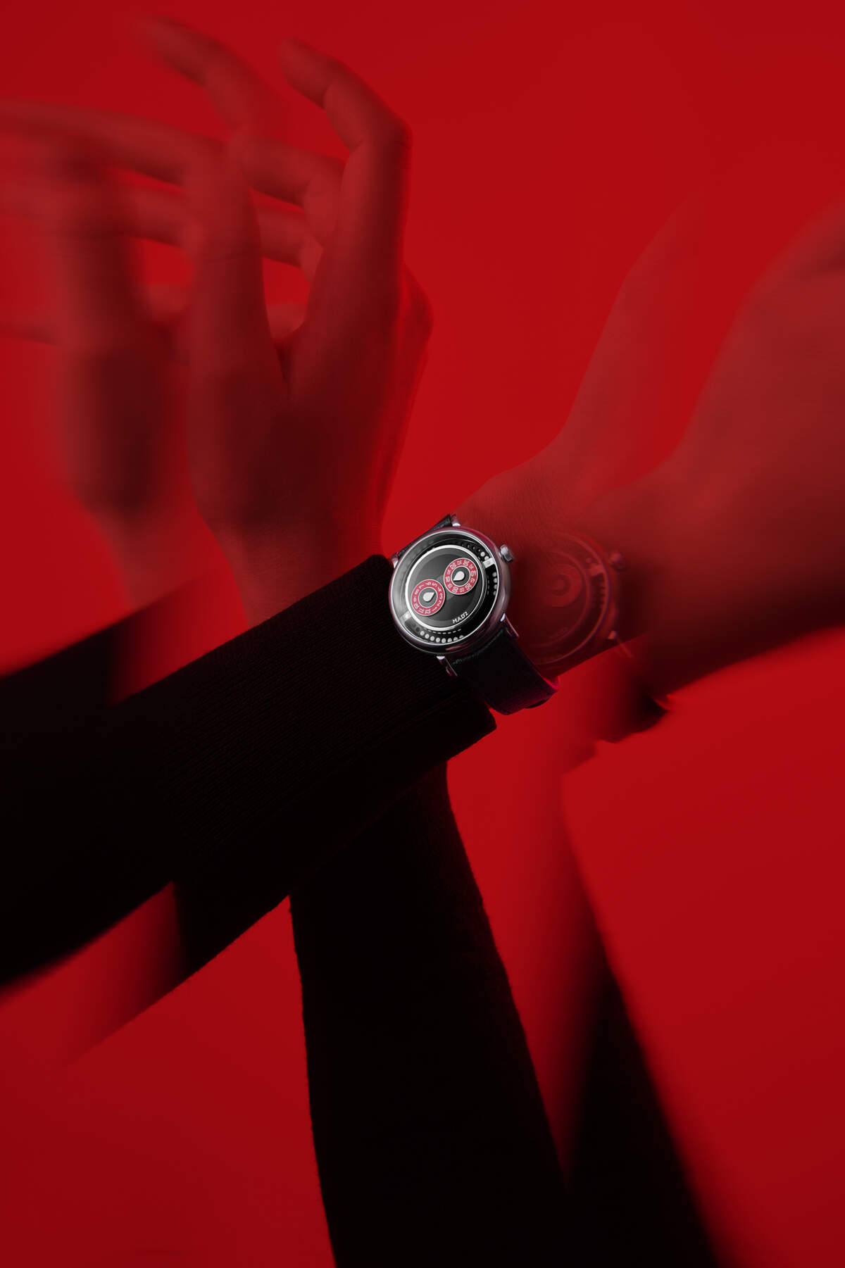 MB&F M.A.D.2 R&B on a wrist in motion against a deep red backdrop, showing the black grooved dial plate, two red sub-dials, polished steel pebble case and black leather strap