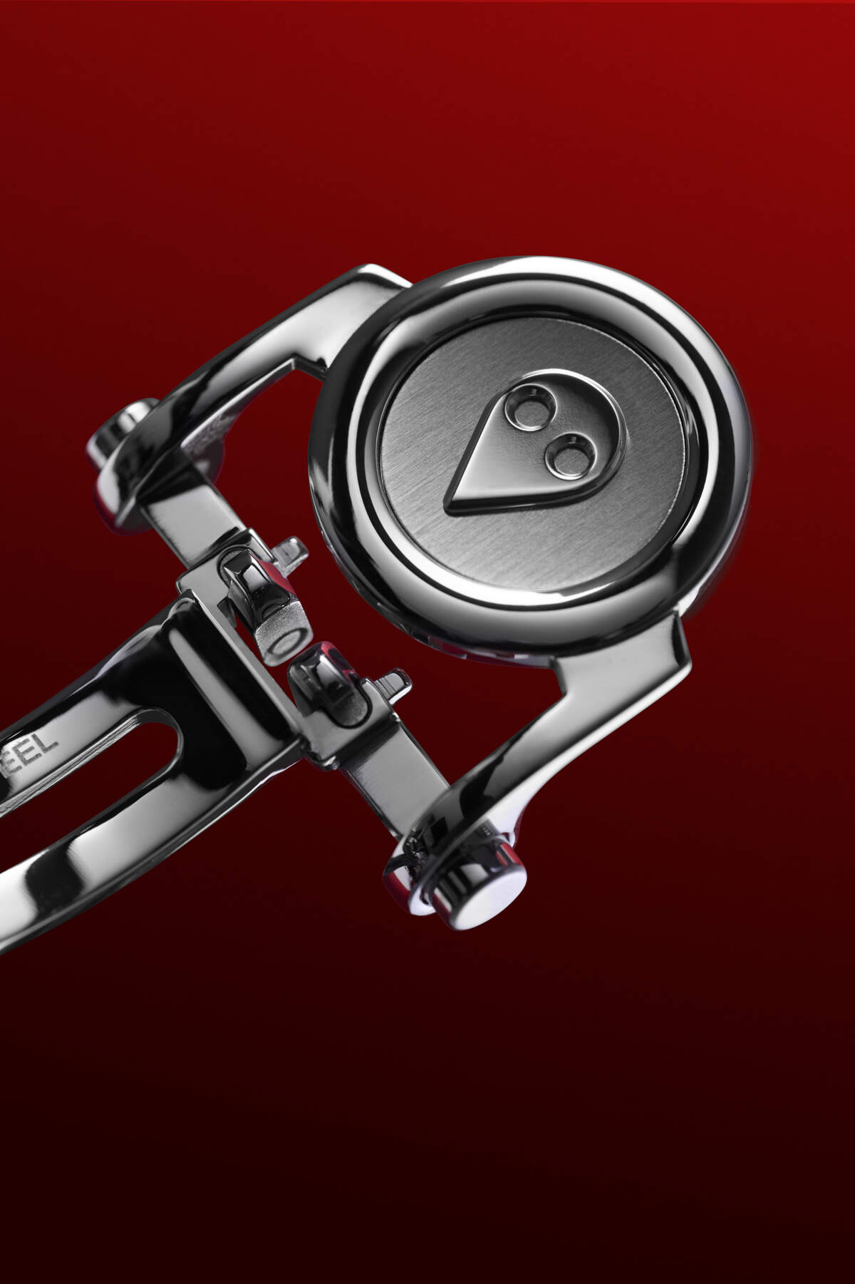 MB&F M.A.D.2 R&B clasp detail showing the polished stainless steel deployant buckle with the embossed alien face, photographed against a deep red background