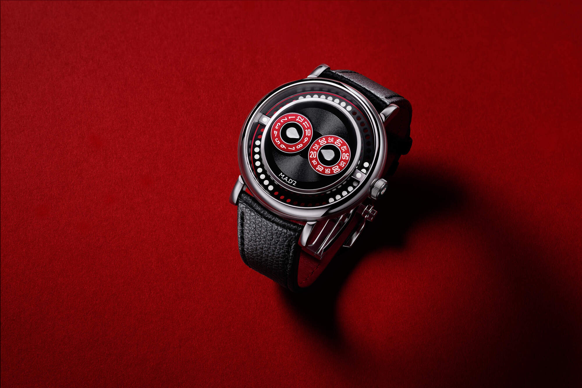 MB&F M.A.D.2 R&B with 42mm polished stainless steel pebble case, black grooved central plate, two raised red sub-dials for jumping hours and trailing minutes, perforated stroboscopic rotor visible beneath, photographed against a deep red background with black leather strap