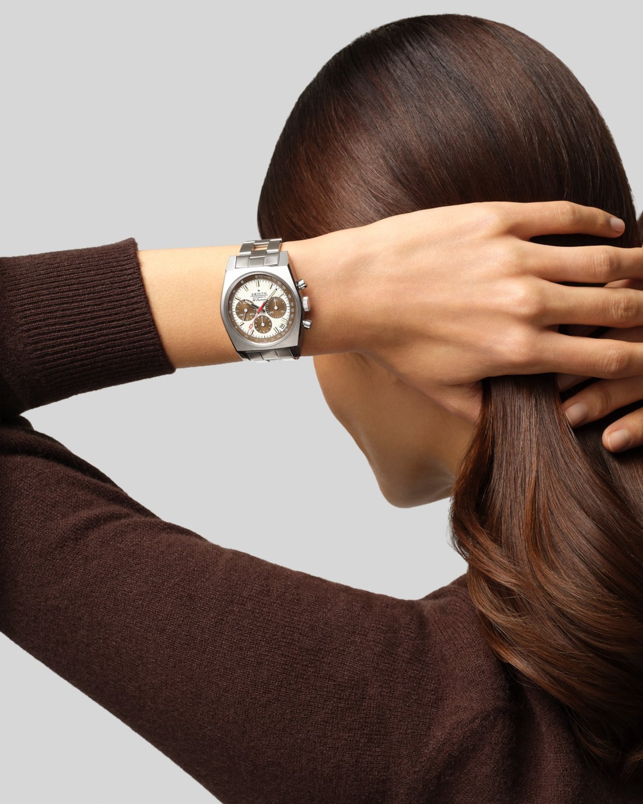 Wrist shot of the Zenith Chronomaster Revival A384 Tropical worn over a brown sweater, showing the white lacquered dial and brown sub-dials of the 37mm tonneau case under natural light, with the steel ladder bracelet visible at the wrist and a hand reaching toward dark hair in the background