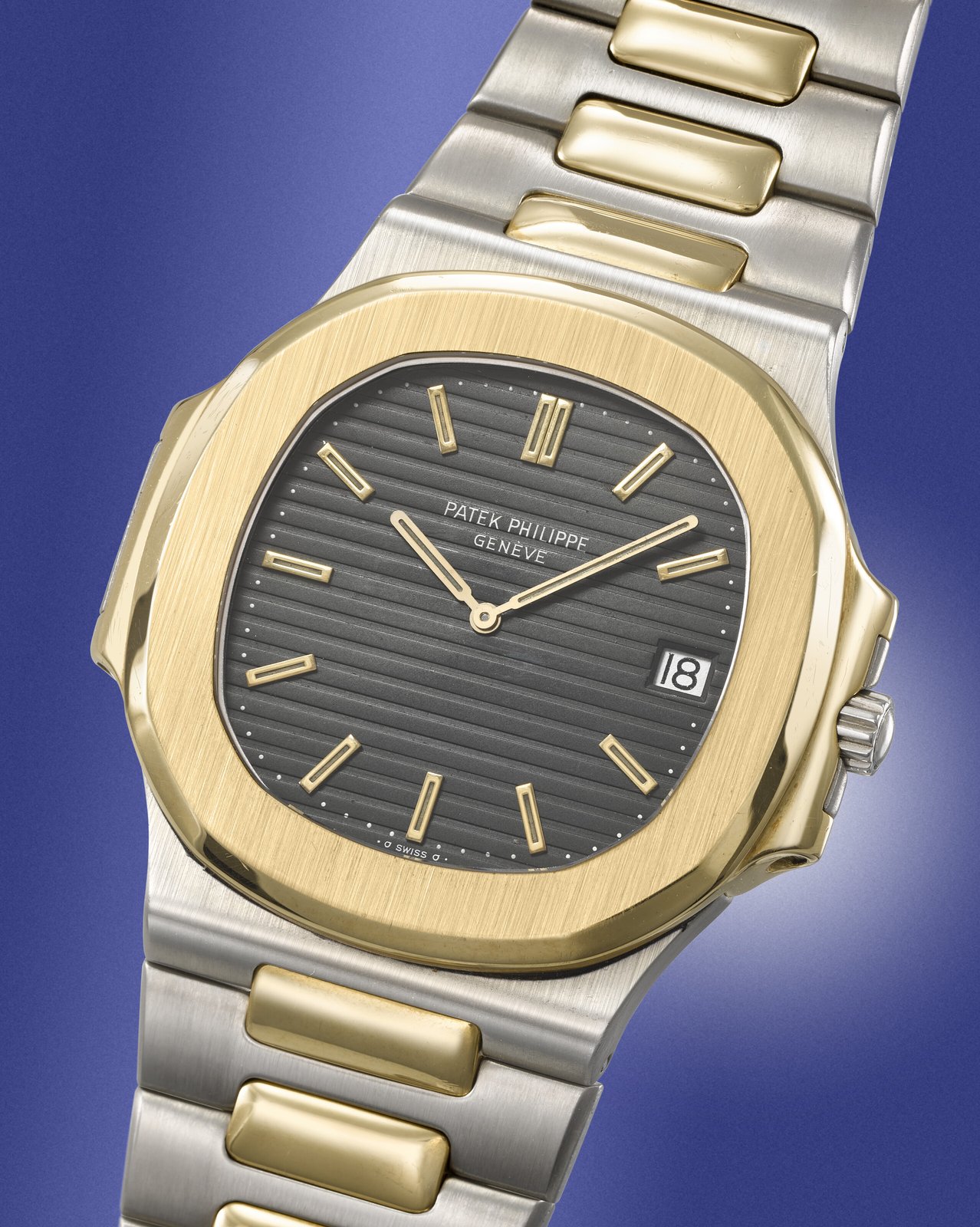 Detail of the matte black horizontally embossed dial of the Patek Philippe Nautilus 3700/1JA, applied yellow gold baton indices, gold dauphine hour and minute hands, date window at three, signed Patek Philippe Genève above the date and Swiss at six, 18-karat yellow gold polished bezel against a blue photographic background