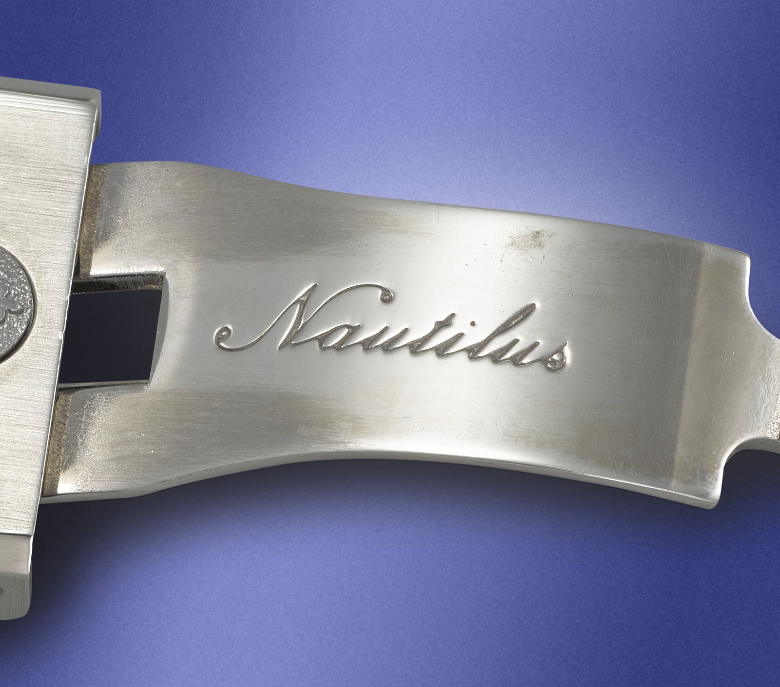 Macro of the folding deployant clasp of the Patek Philippe Nautilus 3700/1JA bracelet showing the original engraved Nautilus signature in cursive script and the Patek Philippe seal above it, brushed steel finish, photographed against a blue background