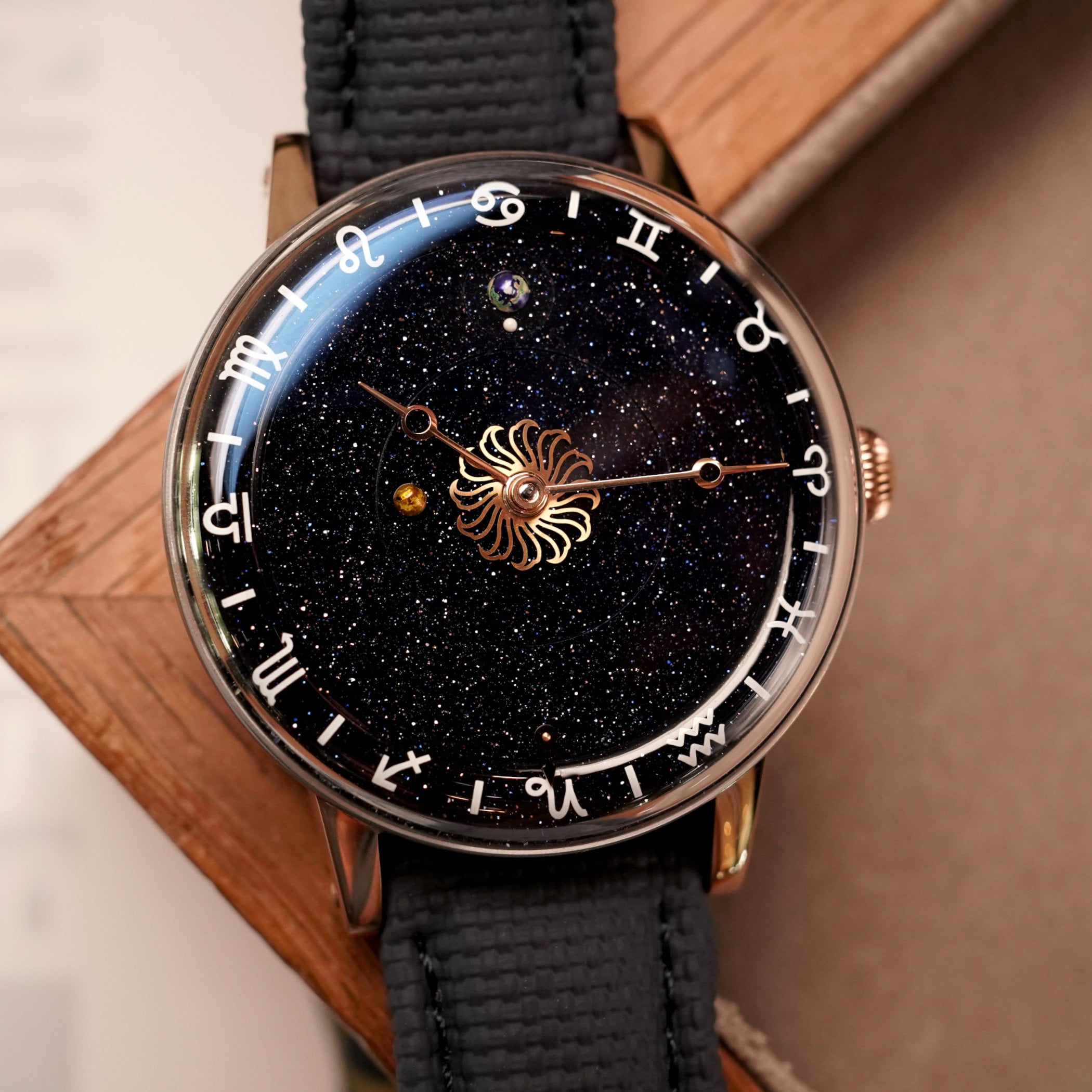 Venus Zodiac rose gold at an angle on a wooden surface, blue aventurine dial with white zodiac flange, onion crown at 3 o'clock, Earth and Moon disc near the top of the dial