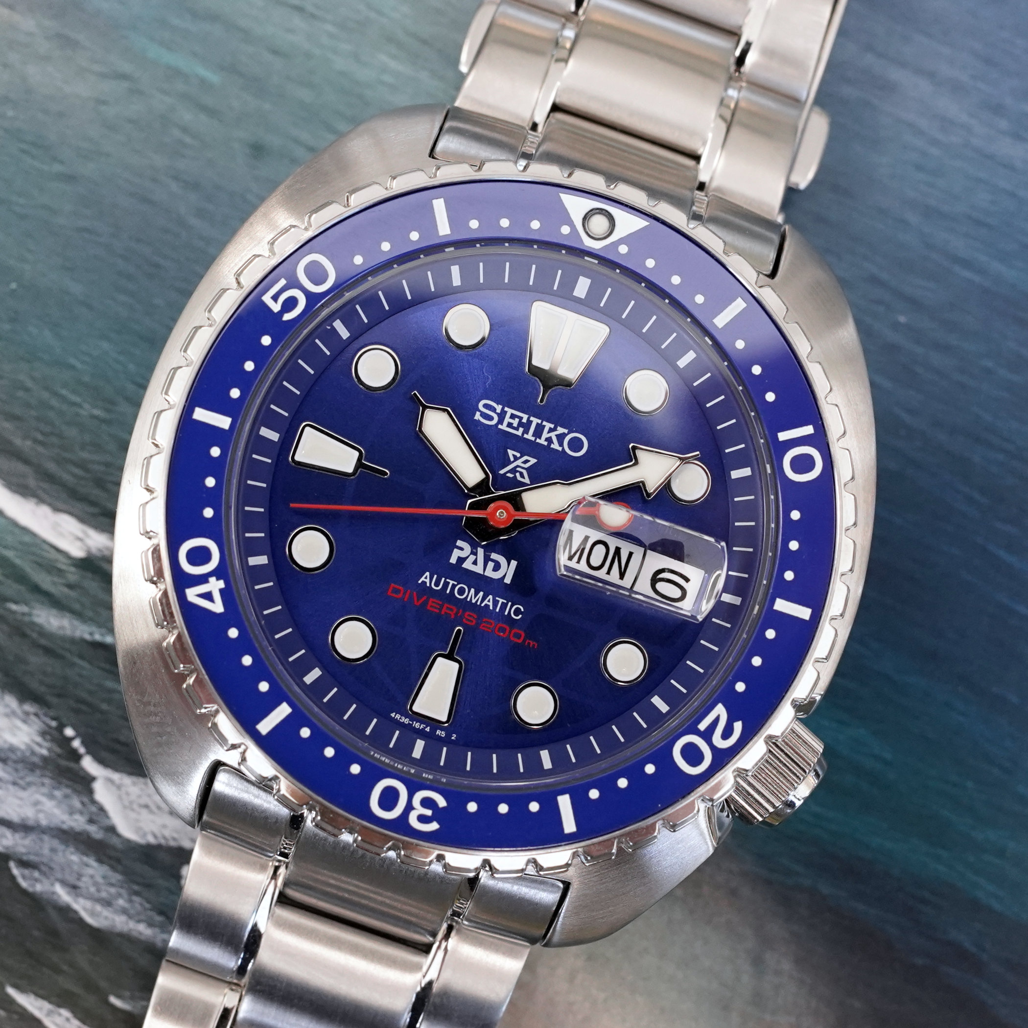 Angled studio macro of the HBB002 electric blue dial, PADI logo and red Diver's 200m text below the centre, blue rotating bezel and day-date window at three, against an ocean background