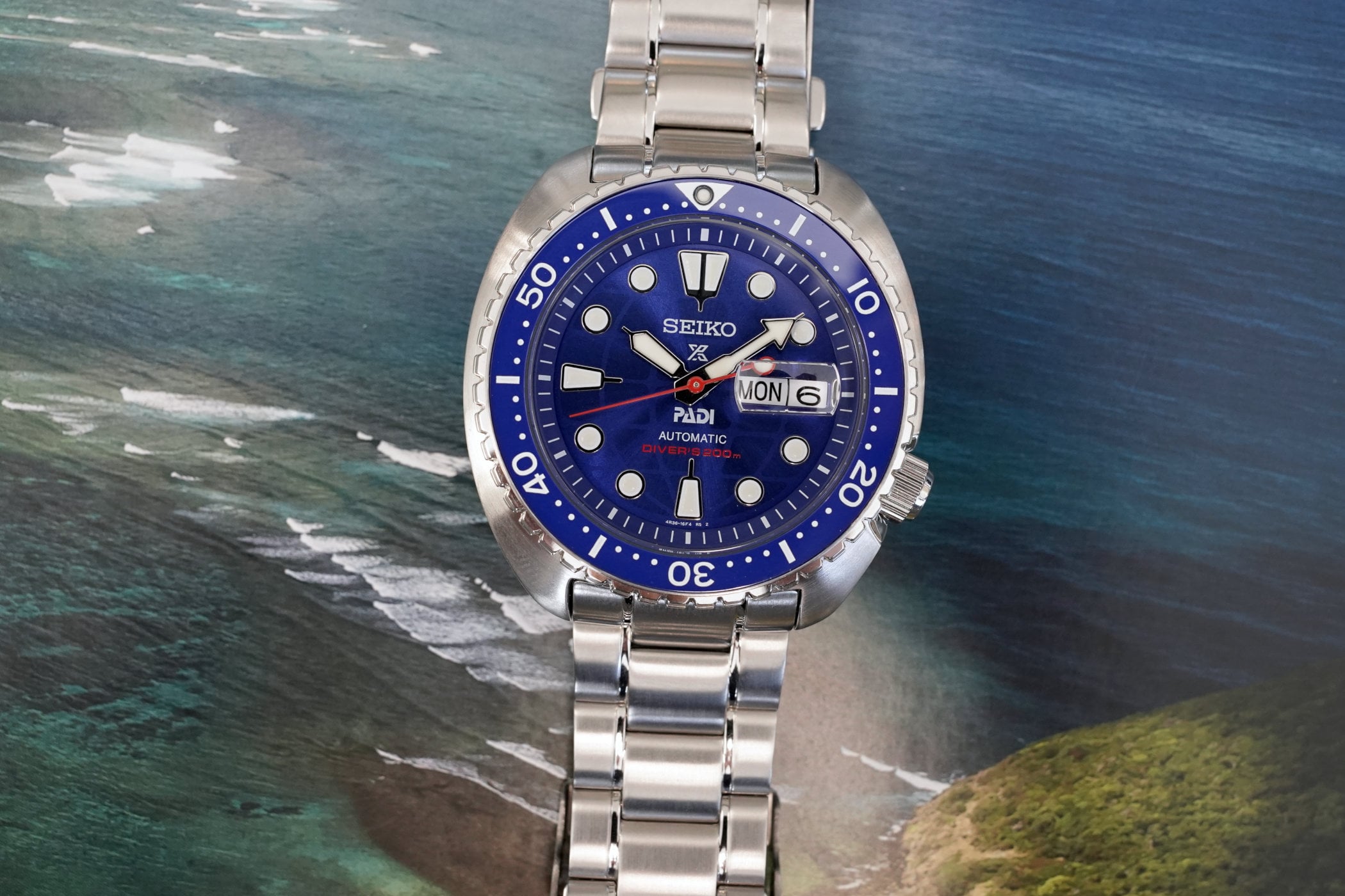 Seiko Prospex PADI 60th Anniversary Turtle reference HBB002, electric blue dial with the PADI logo and red Diver's 200m text above six o'clock, blue rotating bezel, day-date window at three, on a stainless steel bracelet against a coastline