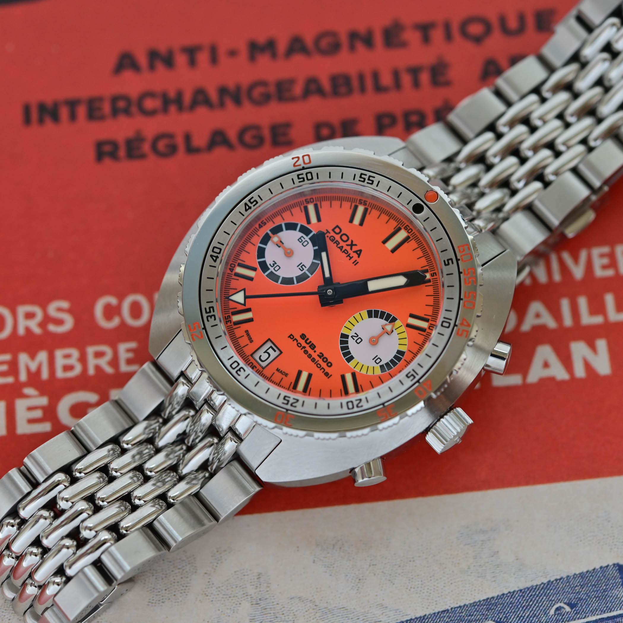 Doxa SUB 200 T.Graph II Professional in orange on a steel beads-of-rice bracelet, bicompax dial with running seconds at 9 and a 30-minute counter at 3, resting on a vintage Doxa brochure