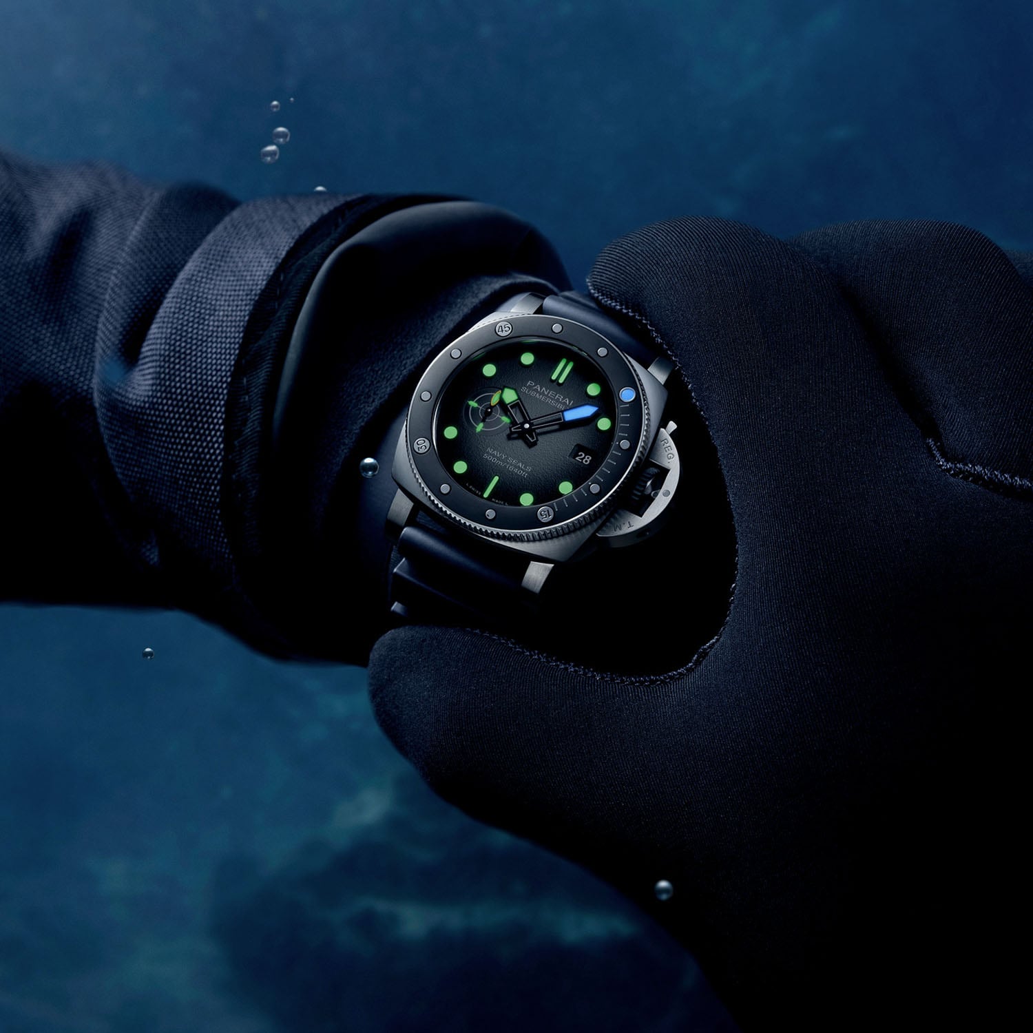 The Panerai Submersible Navy SEALs PAM01738 on the wrist of a wetsuit-gloved diver underwater, the dial glowing green with a blue minute hand and a blue dot at noon on the black ceramic bezel