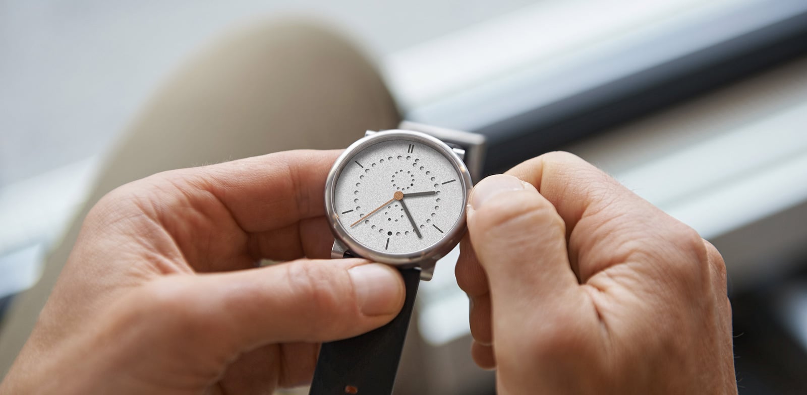 Anno Sandblasted held in two hands with the titanium crown pulled for setting, the full sandblasted grey dial with perforated calendar rings visible, on a black Ecopell leather strap