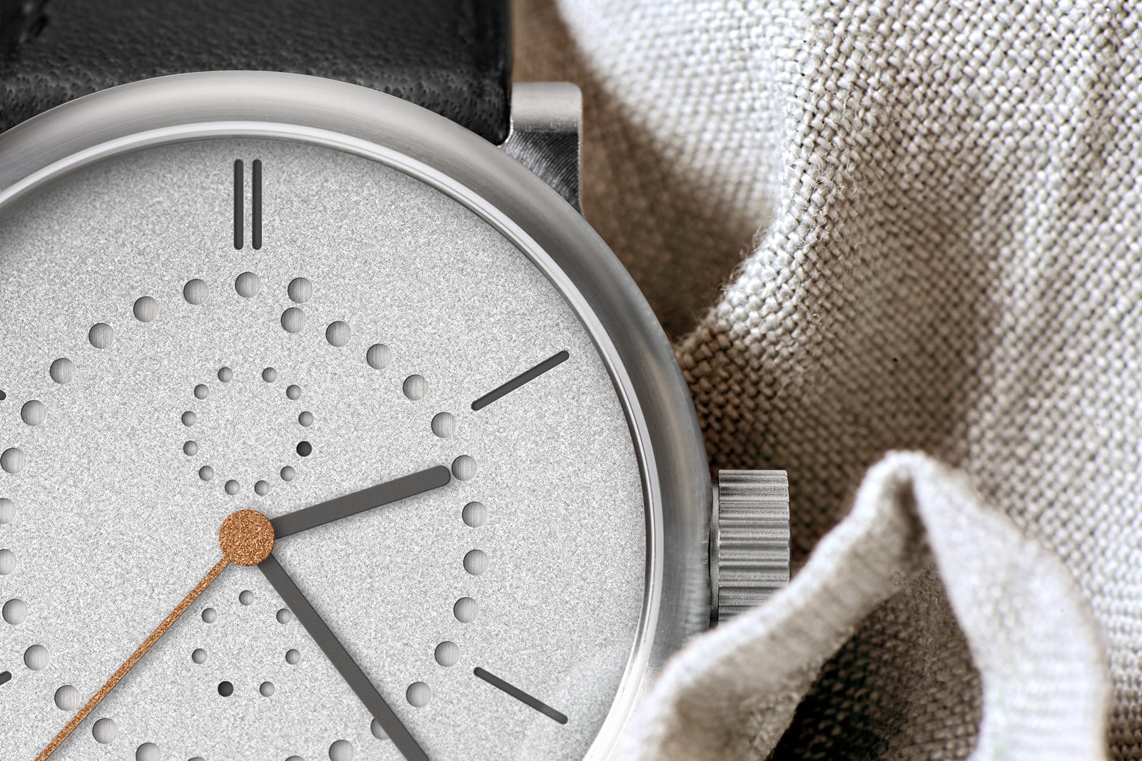 Macro of the Ochs und Junior Anno Sandblasted dial, sandblasted rhodium-plated surface with the perforated date ring at the perimeter and month holes near the centre, gold-leaf seconds hand, fluted titanium crown and black leather strap against linen