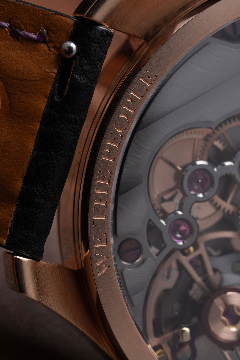Macro of the Liberty 250 case flank, WE THE PEOPLE engraved into the polished 18k rose gold band, with the openworked movement and ruby jewels visible through the front sapphire