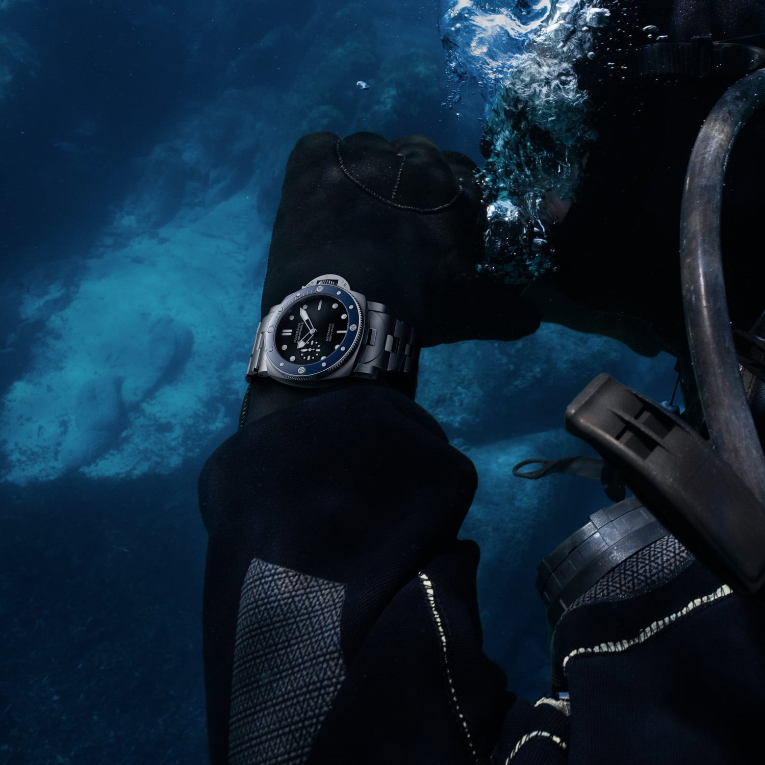 The PAM01756 worn over a black wetsuit sleeve underwater, blue ceramic bezel and luminous dial legible against deep blue water, a second diver and regulator hose in the frame