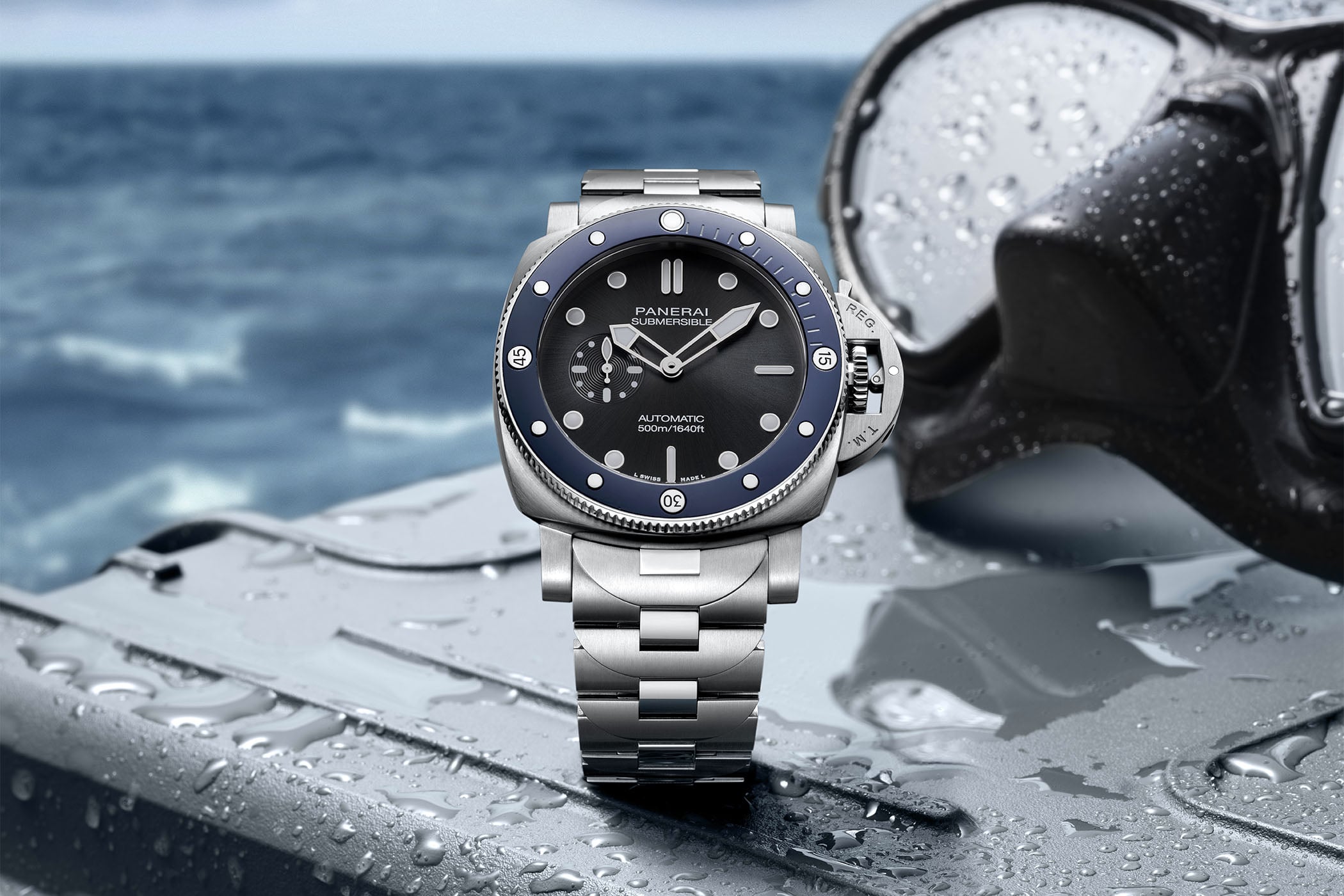 Panerai Submersible PAM01756 on its brushed steel bracelet, 44mm cushion case with blue ceramic unidirectional bezel and black sunbrushed dial reading AUTOMATIC 500m/1640ft, photographed on a wet metal surface with a dive mask and open sea behind