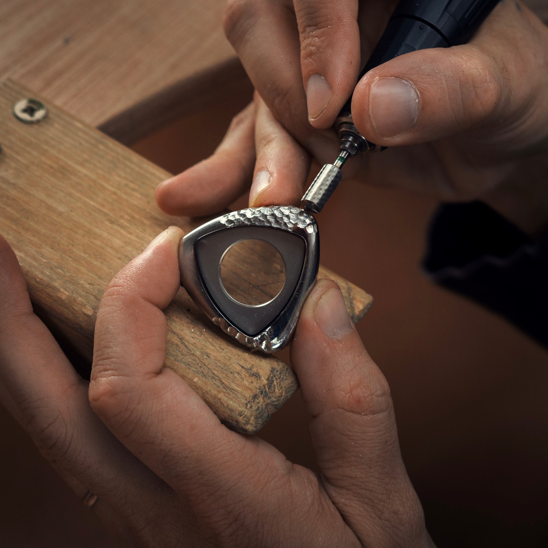 Steven Brunel working the A1 Prehistoric case at the bench with a handheld rotary tool, the triangular steel case held against a wooden block with one flank already pitted and faceted and the rest still smooth