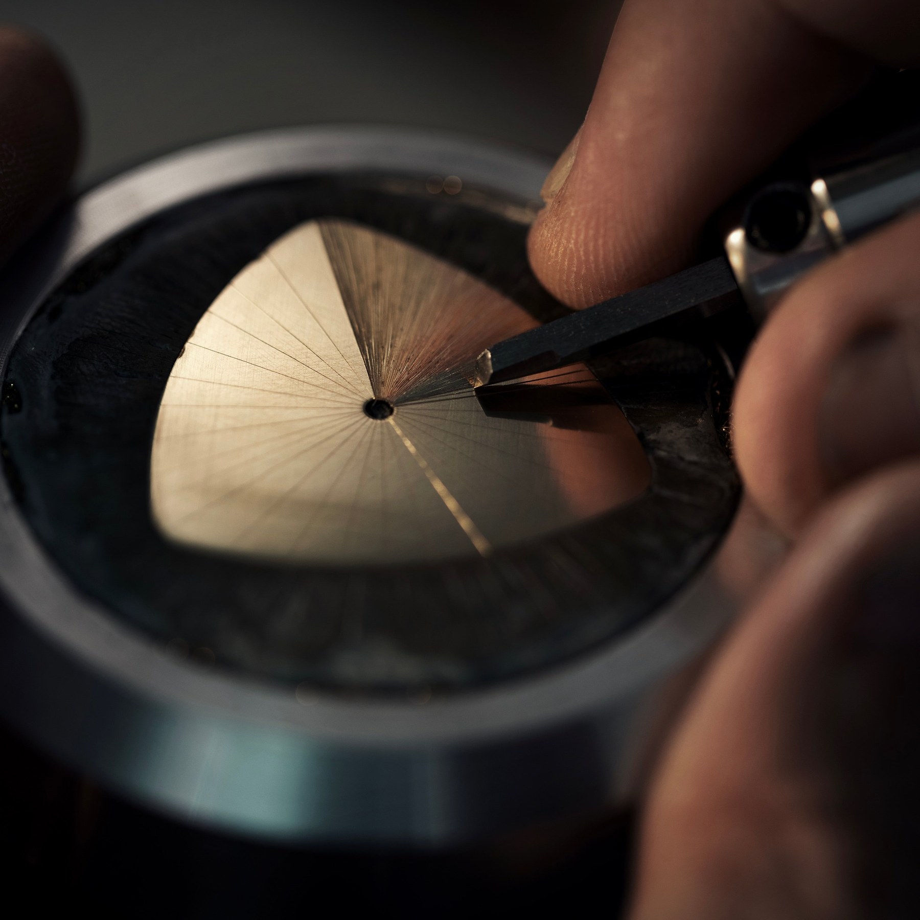 Close-up of the brass A1 Prehistoric dial base held in a fixture while the radiating sunburst lines are cut into it by hand, the bare brass showing gold against the darkened surround