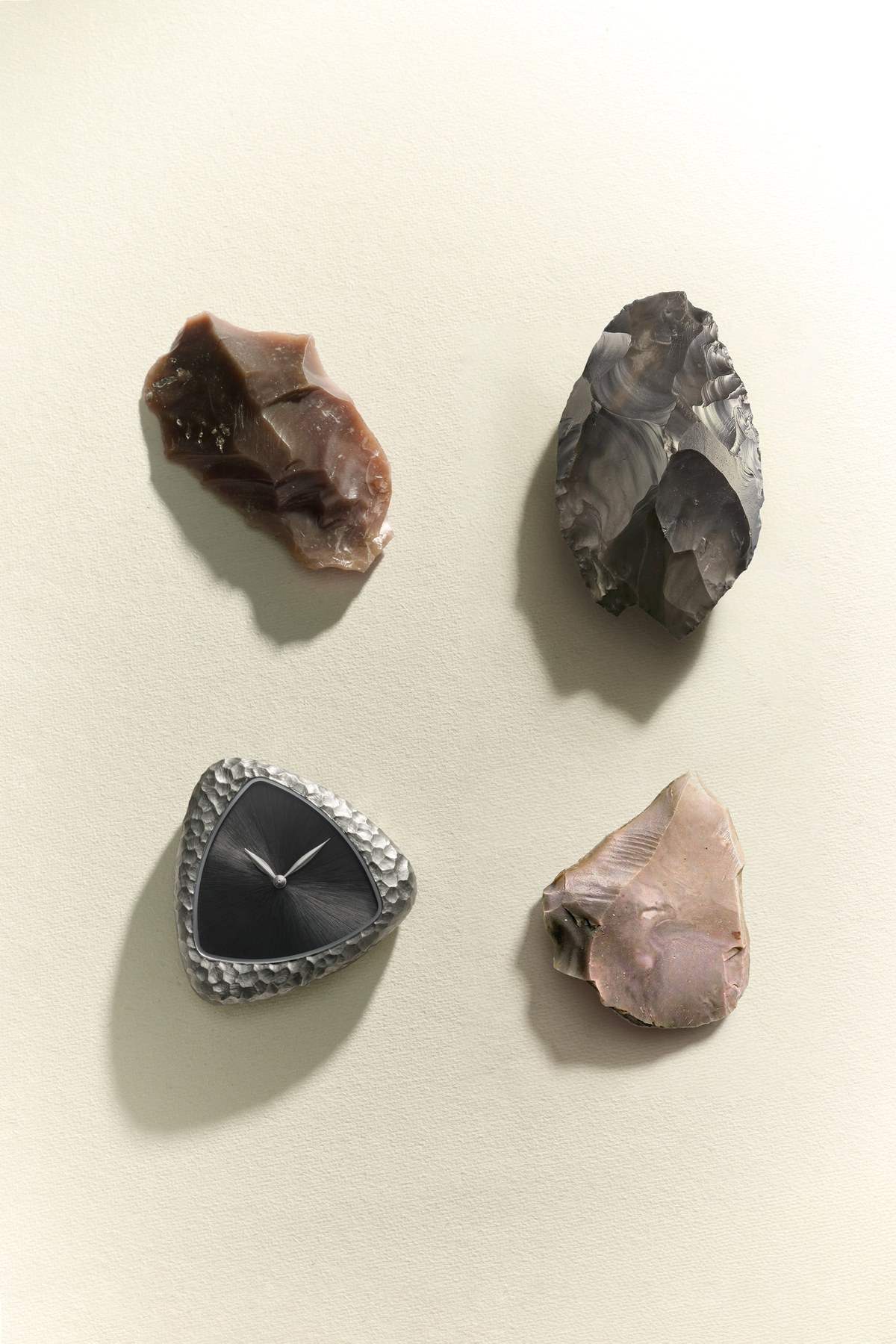 The Anoma A1 Prehistoric watch head laid on a pale ground alongside three knapped flint hand tools, the pitted steel case and dark dial set against the brown and grey stone