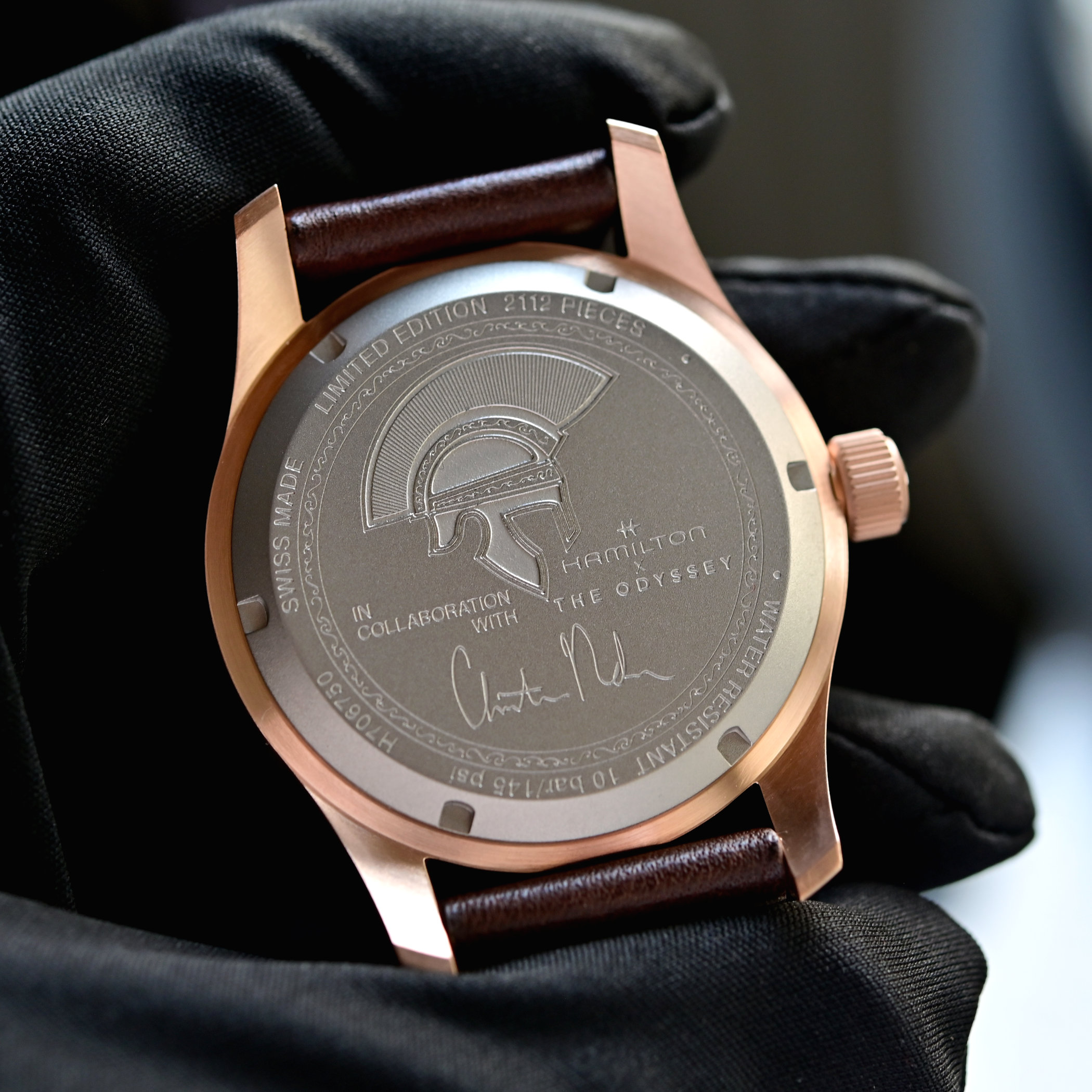 Titanium caseback of the Khaki Field Odyssey held in a gloved hand, engraved with a Greek helmet in profile, the text LIMITED EDITION 2112 PIECES around the top, IN COLLABORATION WITH and HAMILTON X THE ODYSSEY, Christopher Nolan's signature beneath, and WATER RESISTANT 10 bar / 145 psi around the lower edge