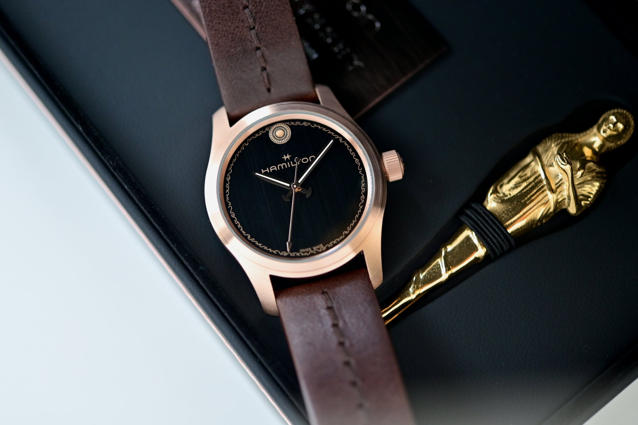 Hamilton Khaki Field Automatic The Odyssey ref. H70675530, 42mm bronze case with a black vertically brushed dial, engraved wave frieze around the dial edge, bronze sword-shaped hands and a spear-shaped seconds hand, on a brown grained leather strap, photographed in its presentation box beside the gold replica of Athena's pin
