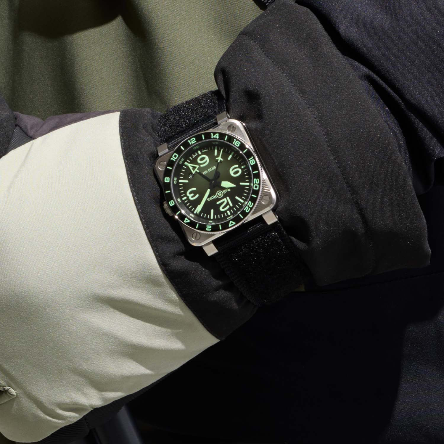Three-quarter view of the BR-03 GMT Green Lum showing the 42mm rounded-square steel case, the four corner screws, the knurled crown and the black polished ceramic bezel insert, on a black rubber strap