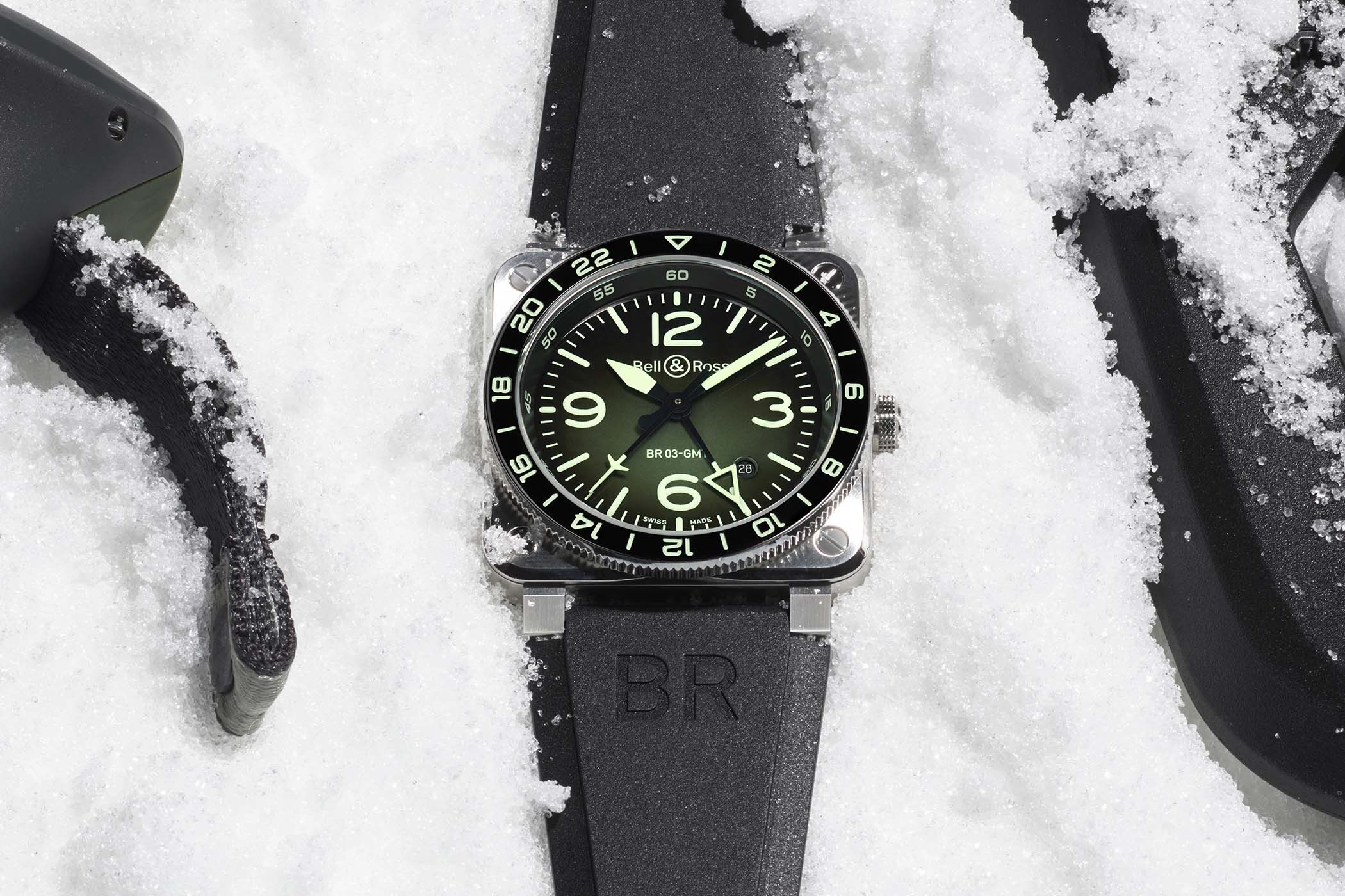 Bell & Ross BR-03 GMT Green Lum resting on snow, a green-to-black gradient dial with pale green Super-LumiNova numerals and markers under a black bezel with a 24-hour scale, on a black rubber strap embossed BR