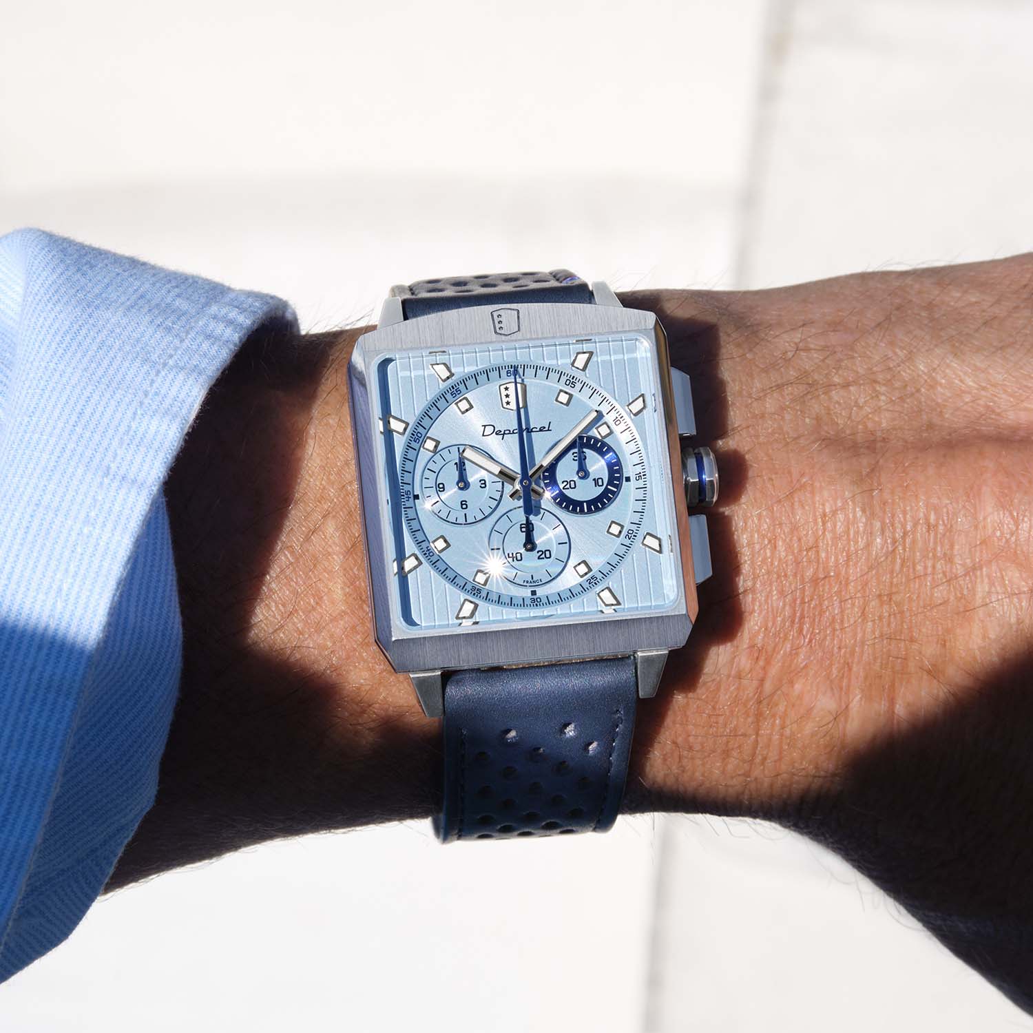 The Depancel Serie-R Ice Blue worn on a wrist in sunlight, square steel case and ice-blue dial with the midnight-blue 30-minute counter at three, on a dark blue perforated rally strap beside a light blue shirt cuff