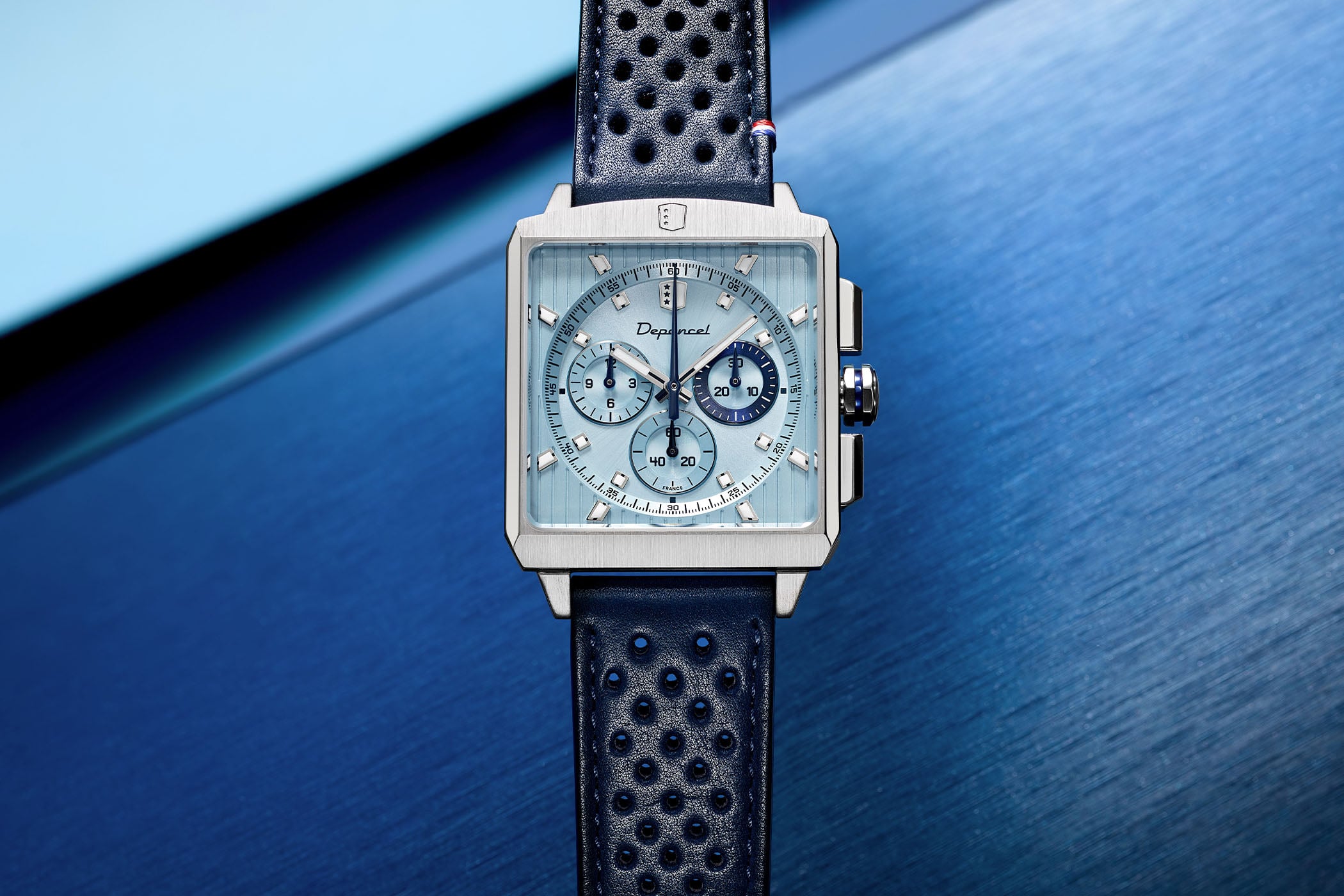 Depancel Serie-R Chronograph Ice Blue, square brushed steel case on a dark blue perforated rally leather strap, ice-blue dial with vertical etched lines, three sub-dials and a midnight-blue 30-minute counter at three o'clock, photographed against a blue background