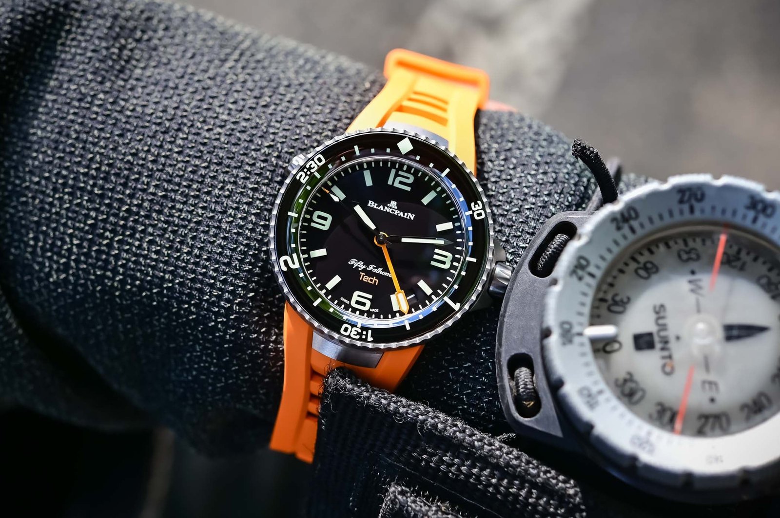Blancpain Fifty Fathoms Tech Ref. 5019A in satin titanium on an orange rubber strap, Absolute Black dial with green and blue luminescence and an orange central three-hour dive hand, worn on the wrist beside a Suunto dive compass
