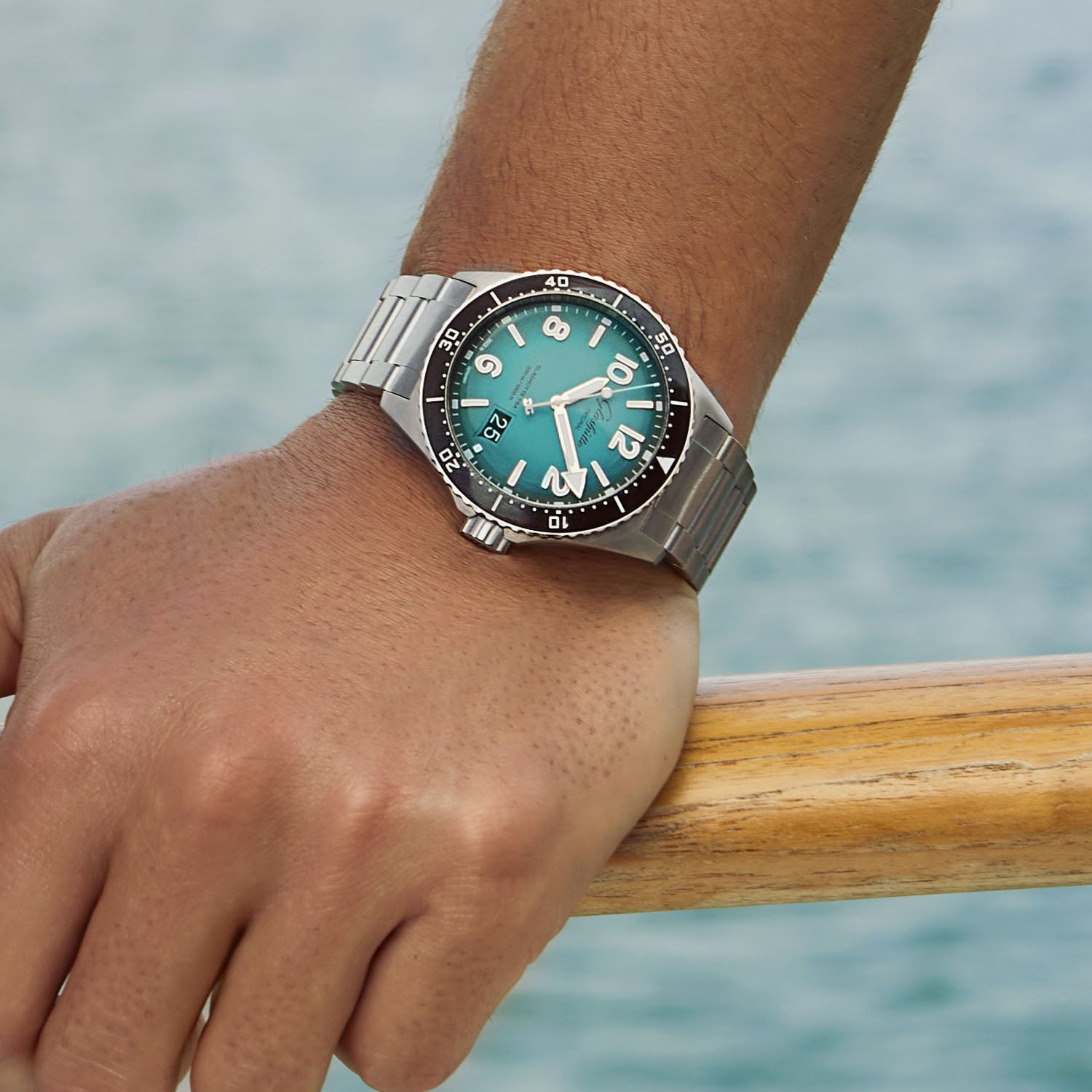The Glashütte Original SeaQ Northern Tide on a steel bracelet worn on the wrist and resting on a wooden rail above water, turquoise-to-black dégradé dial with date at three o'clock