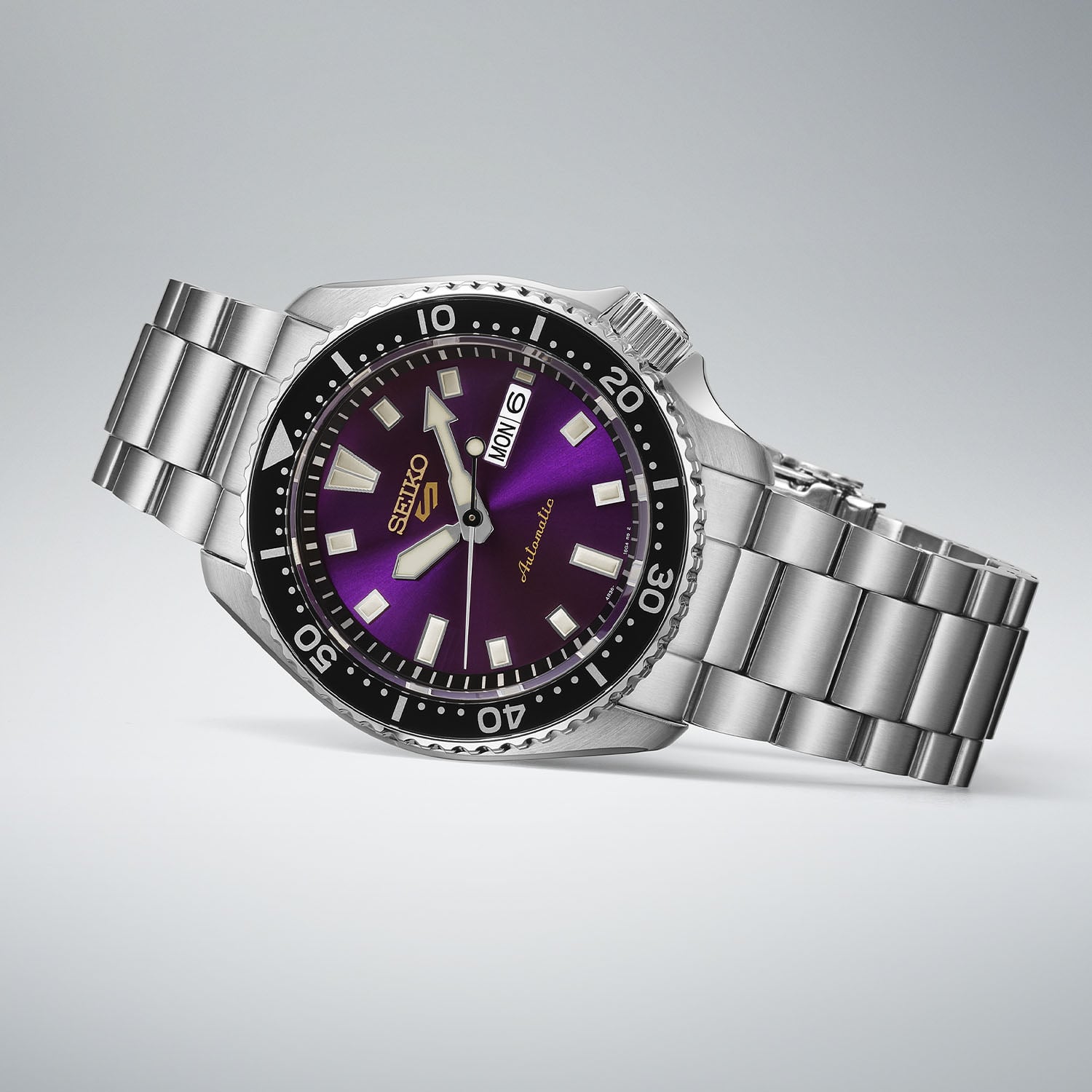 The Seiko 5 Sports SKX HDB003 at a three-quarter angle, deep purple sunray dial with a black rotating bezel, day-date window and gold Seiko 5 logo, on a steel bracelet