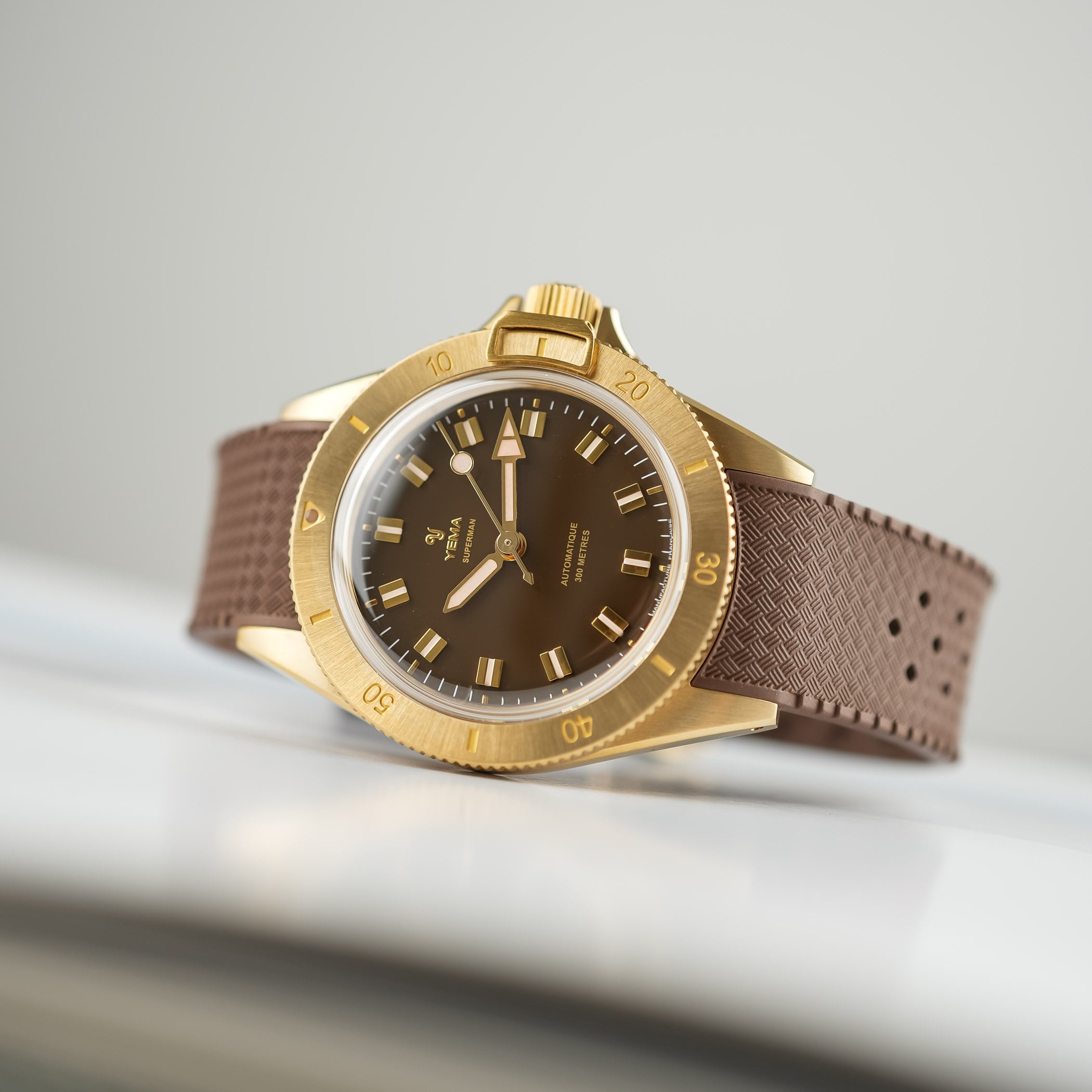 The Yema Superman Bronze CMM.10 in profile, bronze case and radially brushed bronze bezel with the bezel-lock catch, on a brown Tropic-style rubber strap