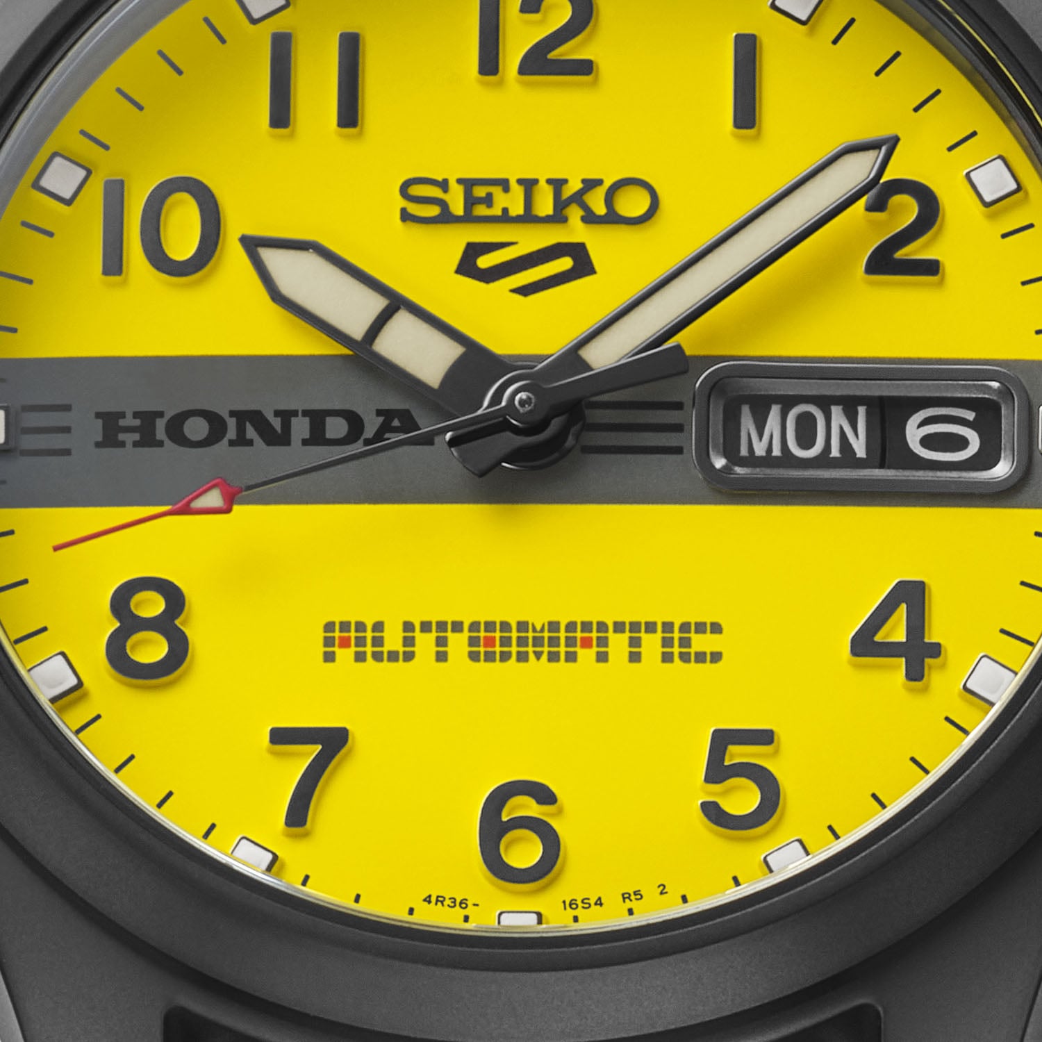 Close-up of the Seiko 5 Sports Motocompo HDB010 yellow dial showing the black Honda bar, the Automatic text in the Motocompo typeface, the day-date at 3 o'clock and the red-tipped seconds hand