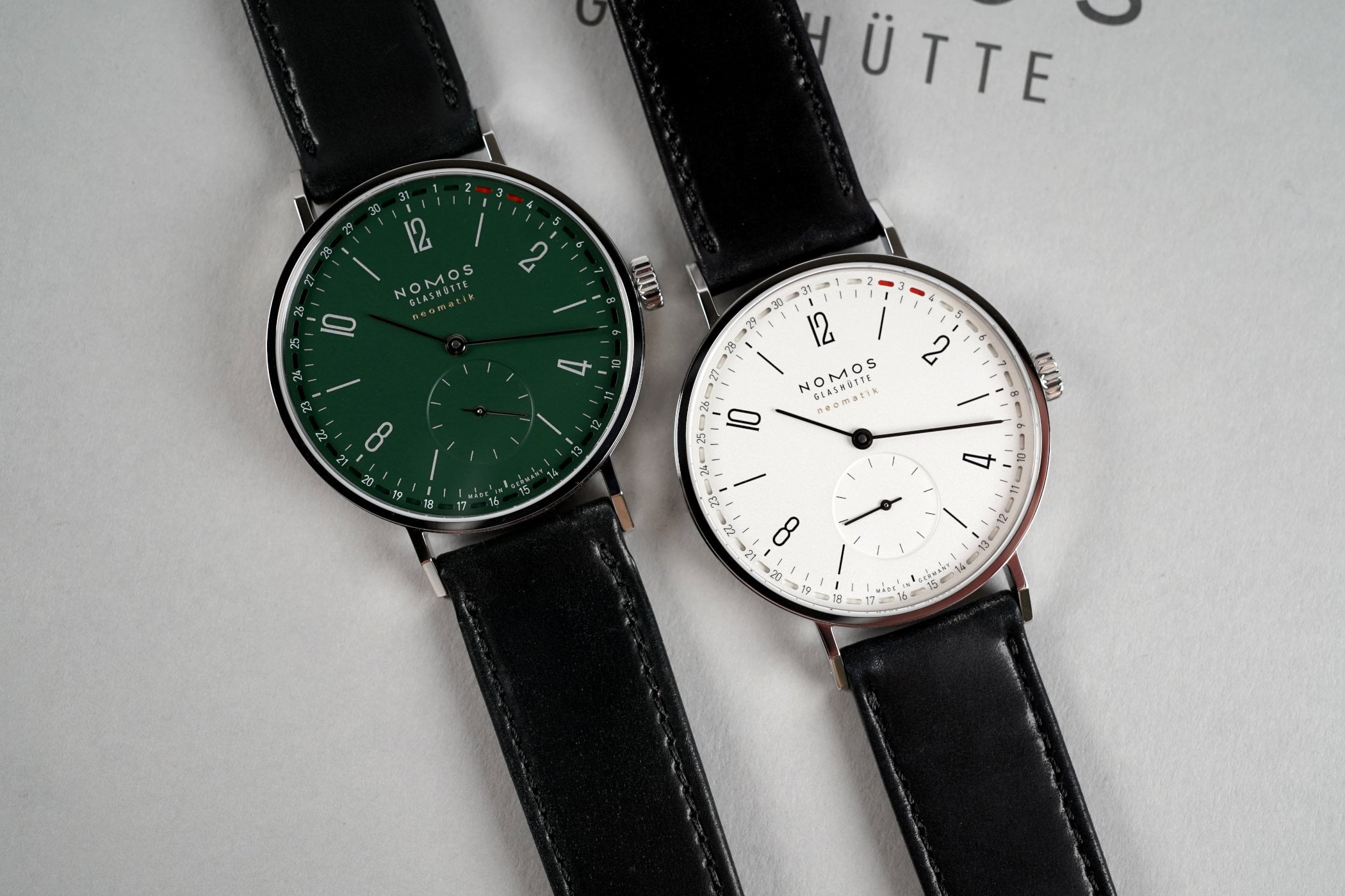 Two Nomos Tangente Neomatik 38 Update watches side by side, a forest-green dial reference 148 on the left and a silver dial reference 147 on the right, both with peripheral date rings marked in red and small seconds at 6, on black leather straps