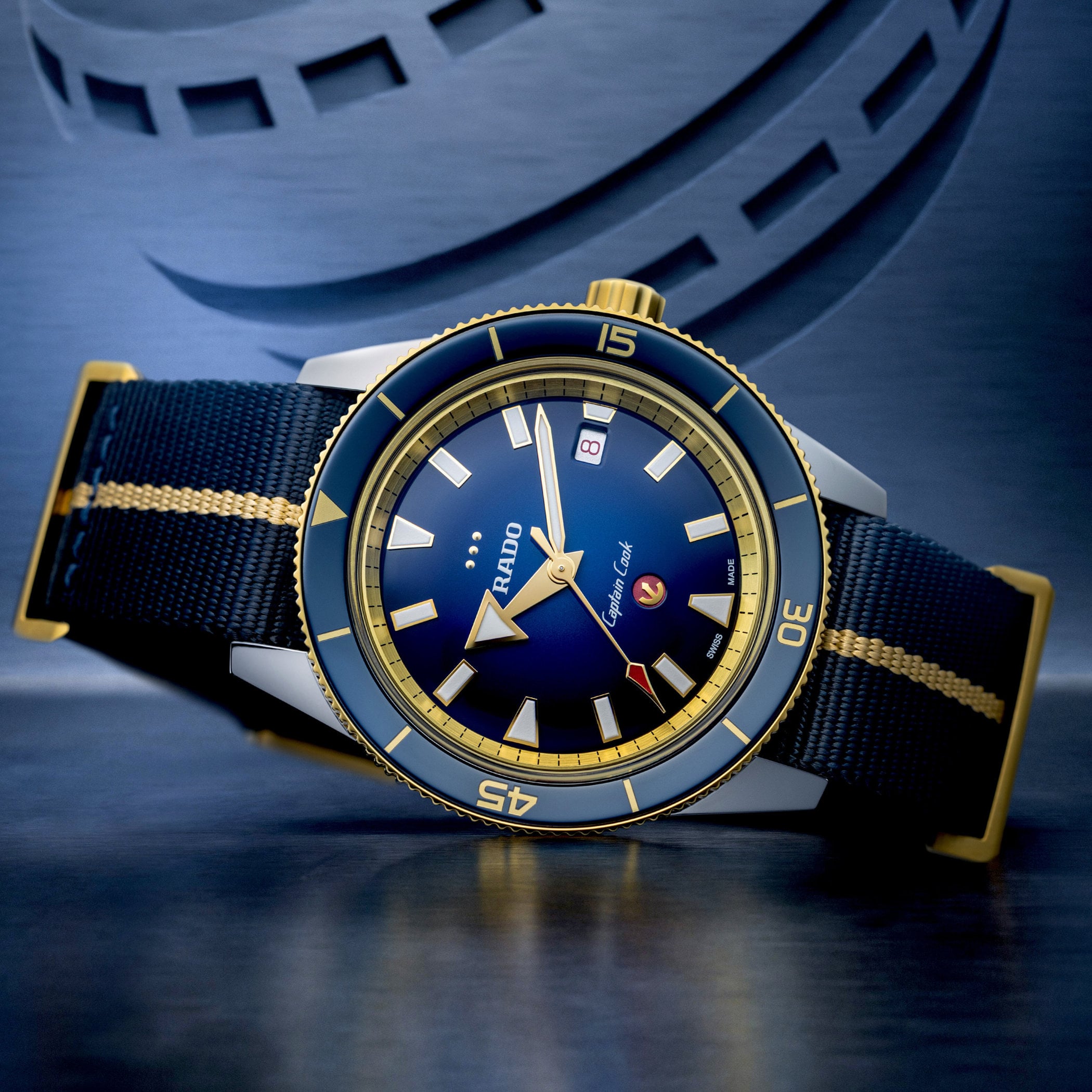 The Rado Captain Cook x European Cricket at an angle on its dark blue and beige NATO-style strap, showing the yellow gold PVD bezel with blue ceramic insert and the blue gradient dial
