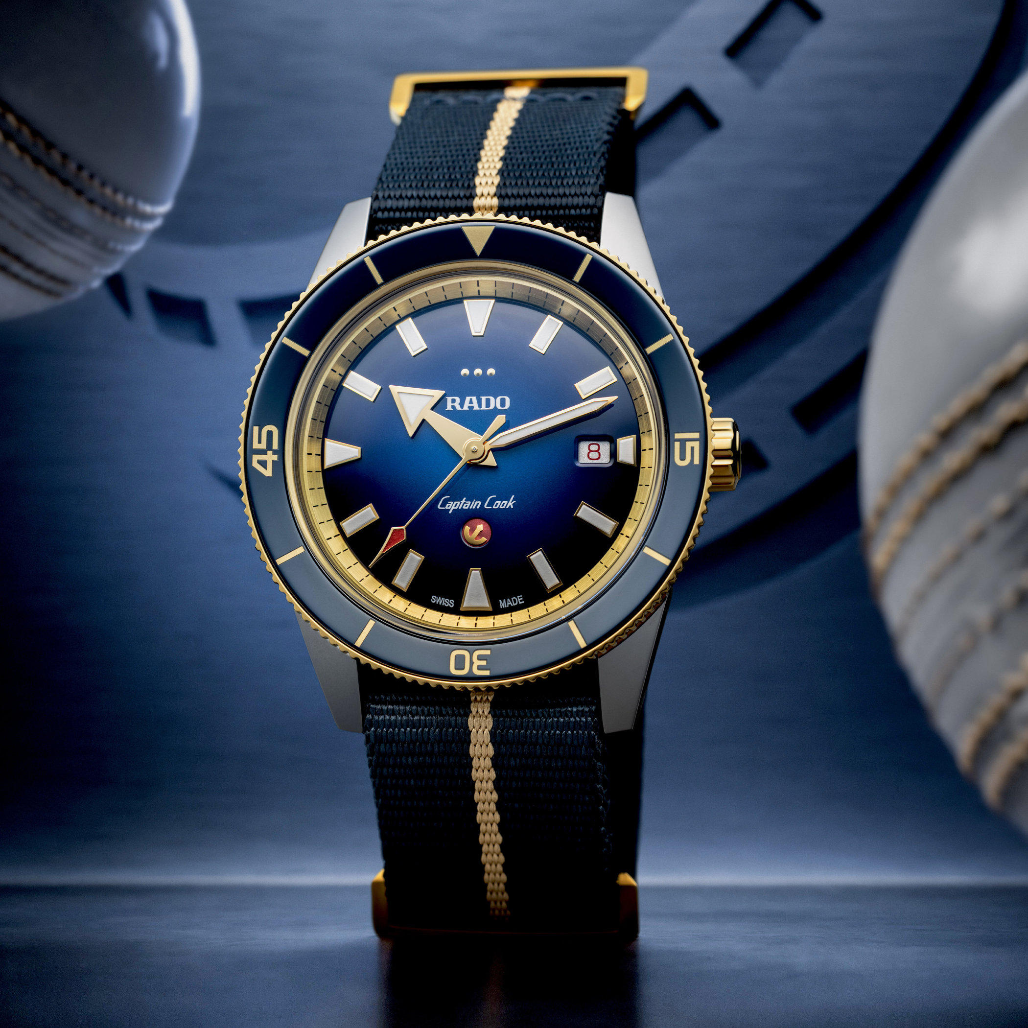 The Rado Captain Cook x European Cricket seen head on, standing on its dark blue and beige NATO-style strap, the blue gradient dial and gold-framed indices in full view with two cricket balls out of focus behind it