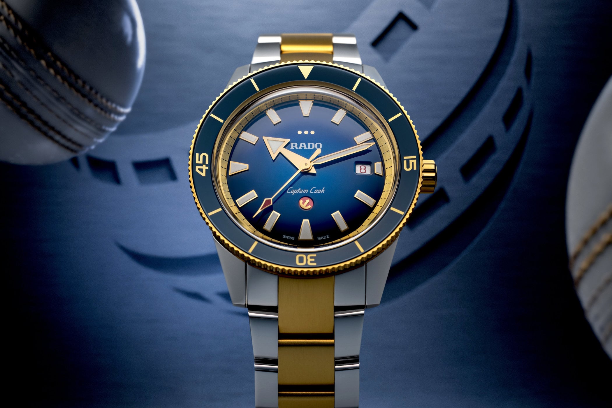 Rado Captain Cook x European Cricket Special Edition, a two-tone steel and yellow gold PVD diver with a blue gradient dial, three gold dots above the Rado logo and a red anchor counterweight, on a two-tone bracelet against a cricket ball