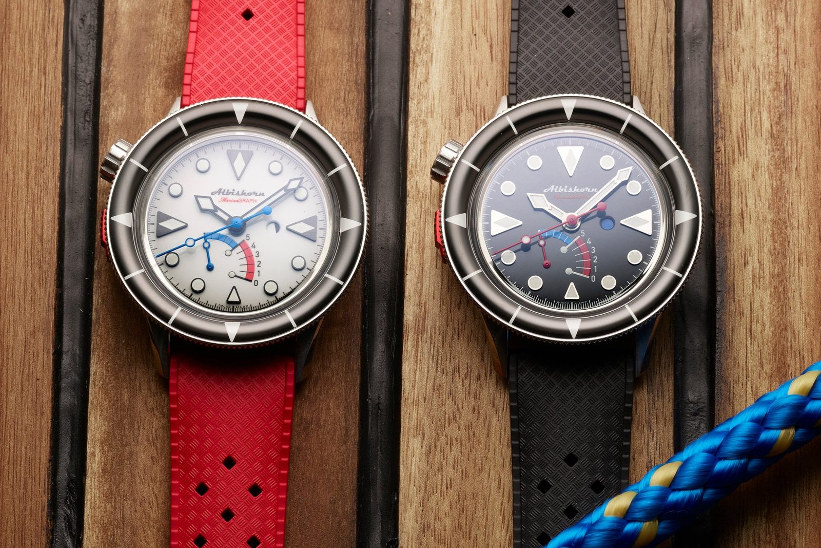 Two Albishorn Marinagraph watches lying on a teak deck, a white lacquered Classic Fumé on a red rubber strap on the left and a sunburst black Classic on a black rubber strap on the right, both with a blue-and-red retrograde countdown arc in the lower dial