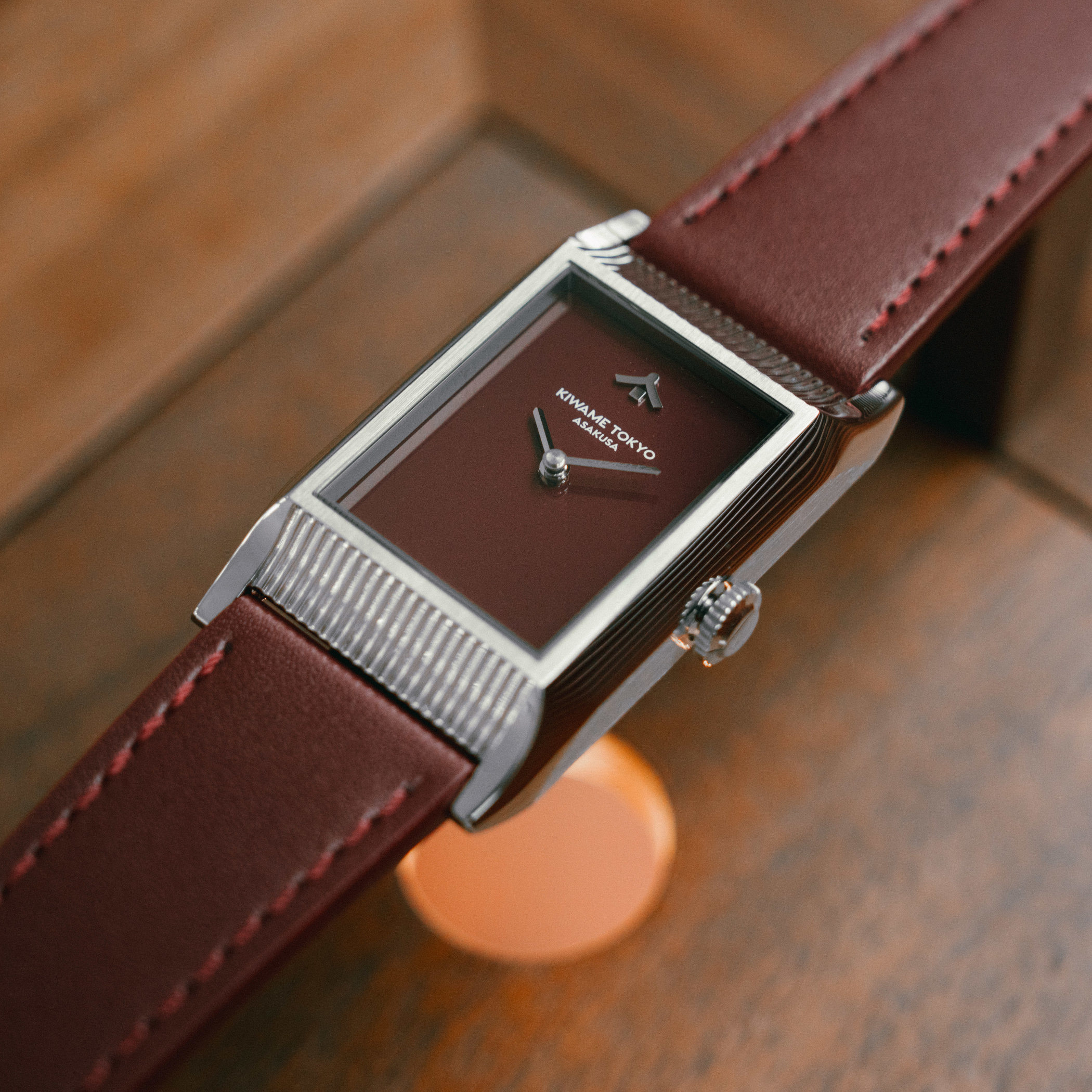 The brown-dial Kiwame Tokyo Yane at an angle on dark wood, its burgundy-brown lacquer dial carrying only a single mune-shaped index at twelve and baton hands, the reeded roof-tile case ends and fluted crown catching the light