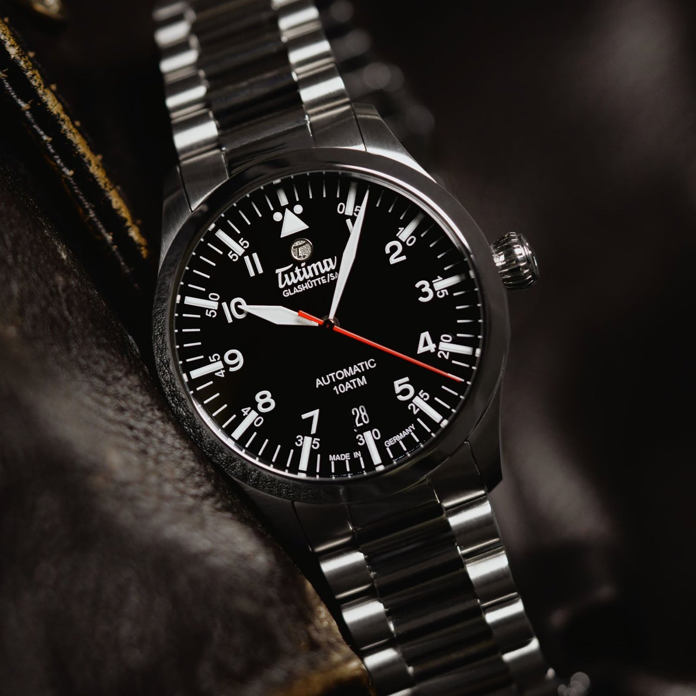 The black-dial Tutima Flieger T5, reference 6109-02, at an angle on its titanium bracelet, resting on leather, showing the domed sapphire crystal, the printed minute track and the fluted screw-down crown