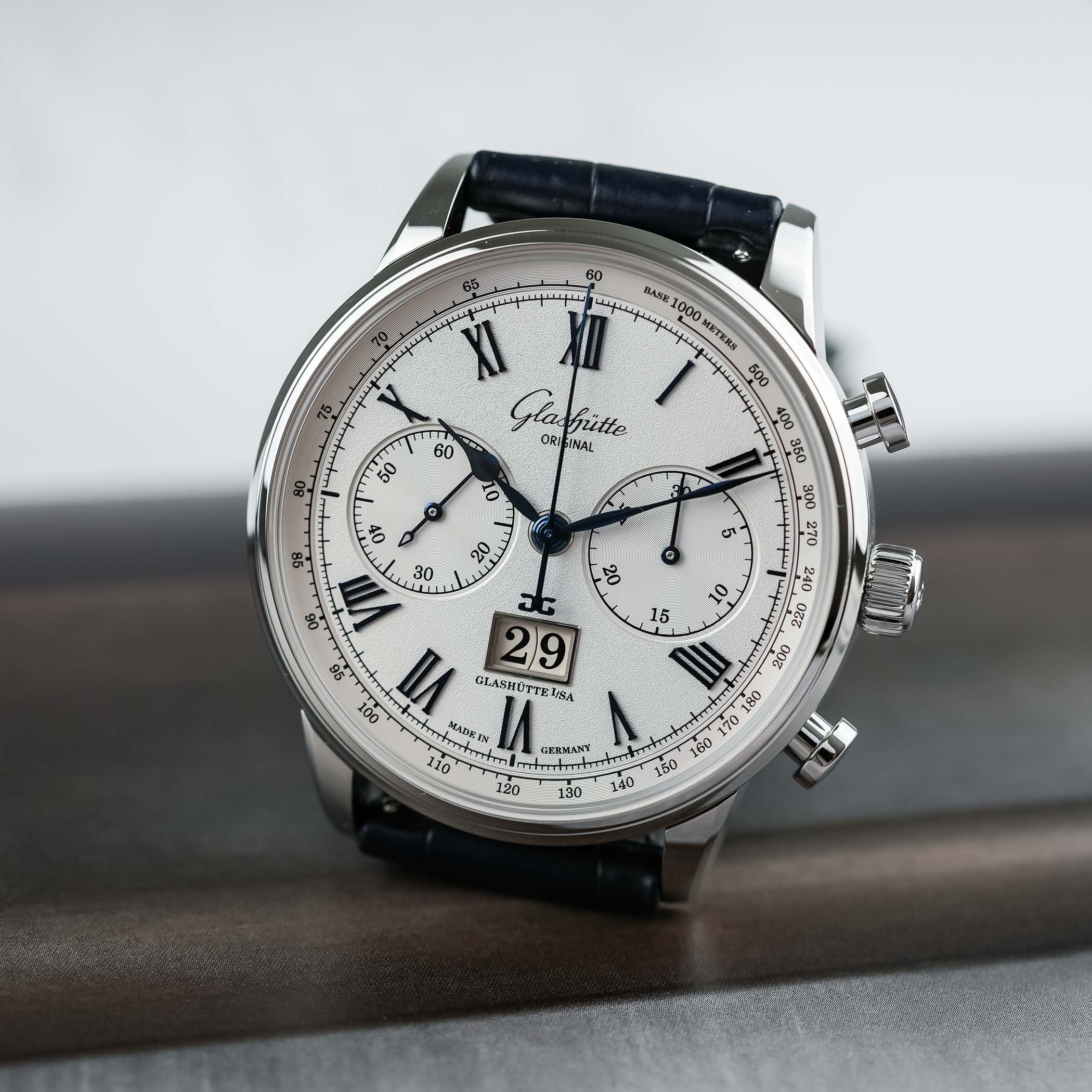 The classic Glashütte Original Senator Chronograph at an angle on a blue alligator strap, its silver-white grainé dial carrying blued solid-gold Roman numerals, blued hands, two recessed vinyl-record registers and a white-gold-framed Panorama Date at six