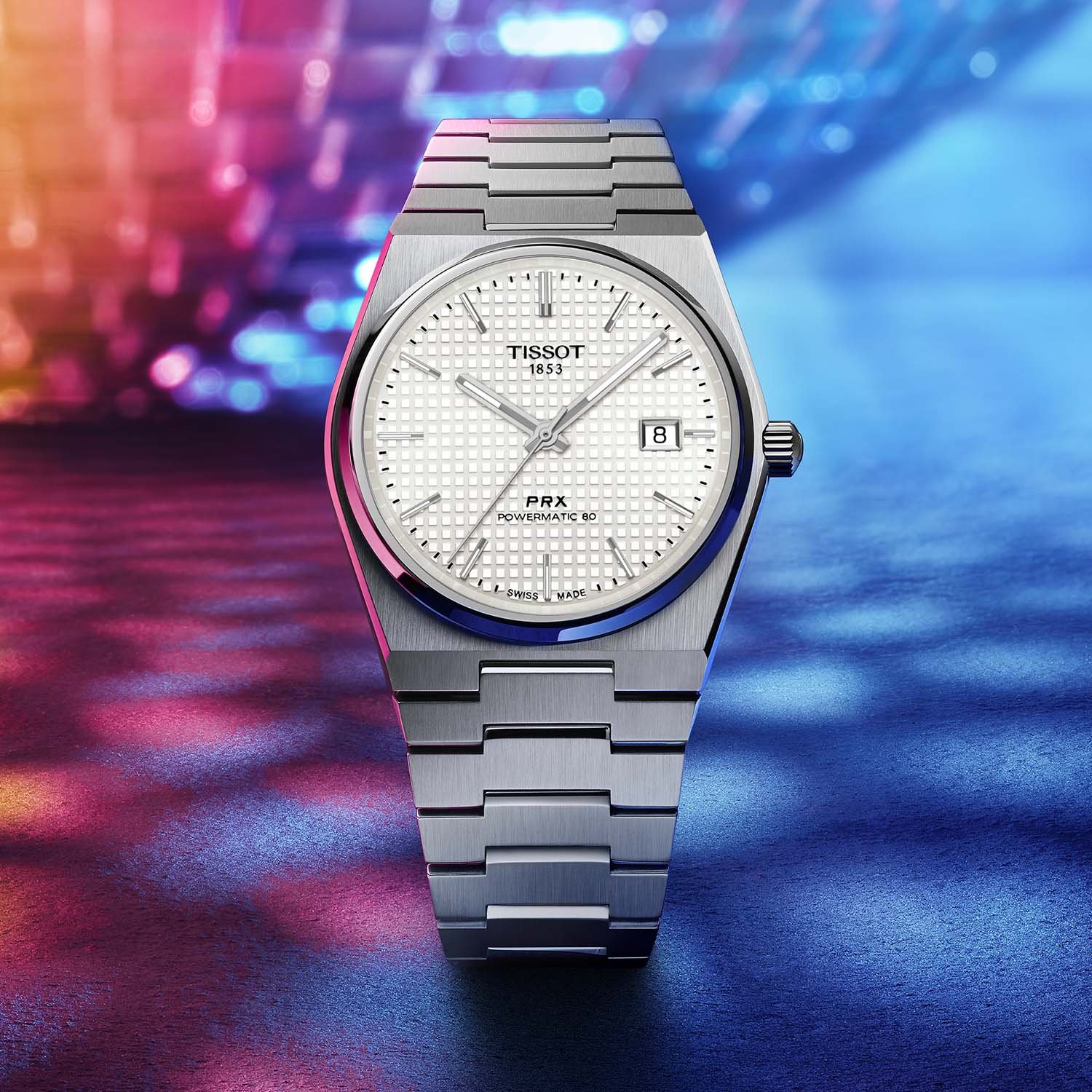 A front view of the Tissot PRX 40mm Powermatic 80 on its steel bracelet against coloured bokeh, the mother-of-pearl waffle dial with applied markers, central seconds and framed date at three