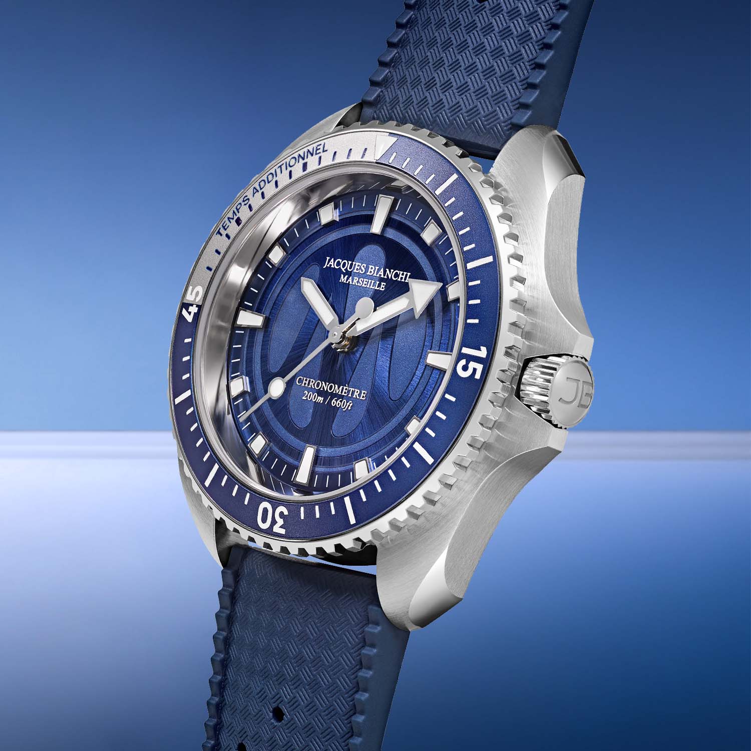 The Jacques Bianchi OM200 at a three-quarter angle on a blue FKM rubber strap, showing the brushed steel case flank, blue 60-click bezel and a crown engraved with the JB monogram
