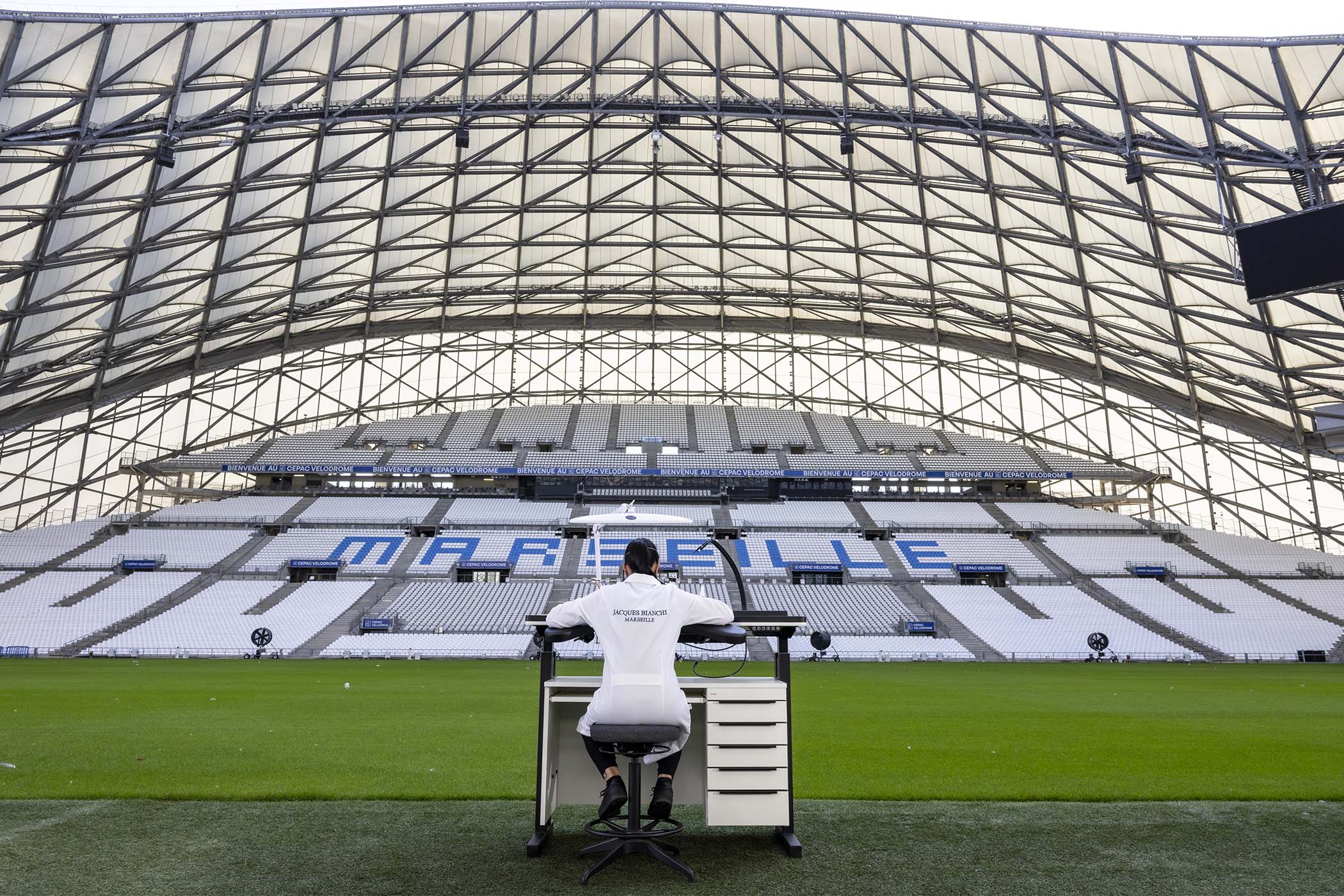 A watchmaker in a Jacques Bianchi Marseille coat working at a bench on the pitch of Marseille's Stade Vélodrome, the empty white stands spelling MARSEILLE behind