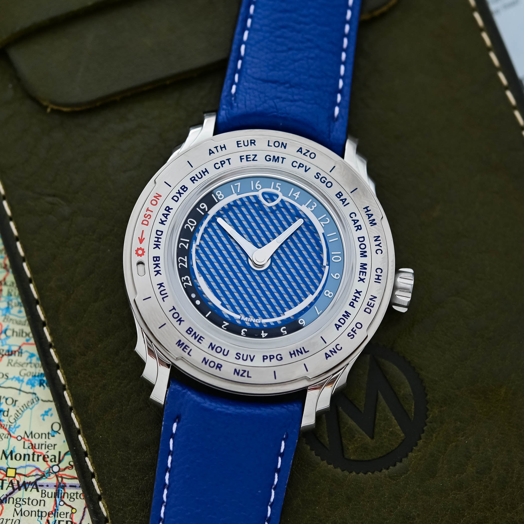 Top-down view of the Ming 57.05 Comet, its rotating 24-hour city ring in blue and black around the striped blue Comet Tail dial with the red DST ON indicator near nine o'clock, on a cobalt-blue strap