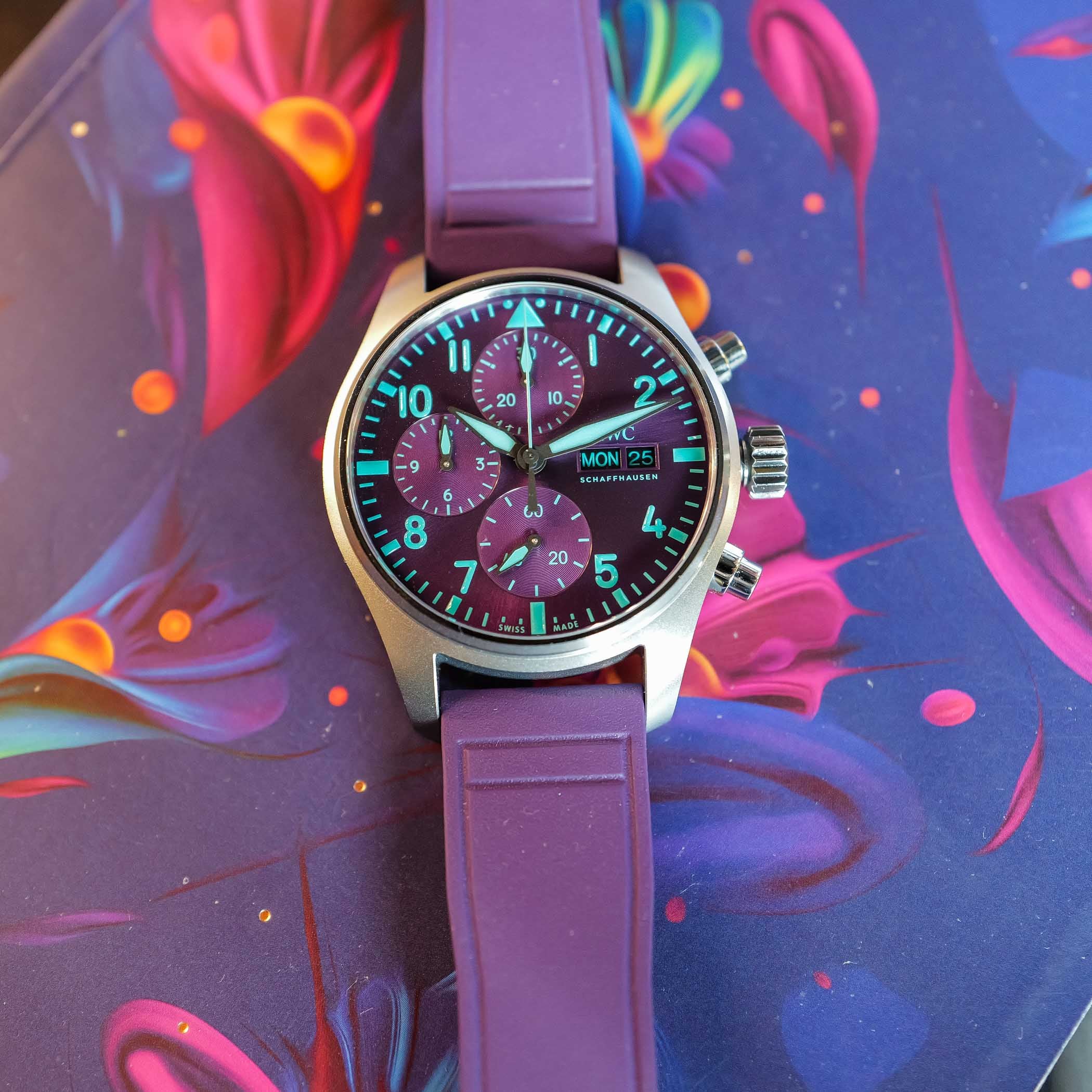 Top-down view of the IWC IW388121 purple dial, its three teal-green chronograph subdials, arrowhead marker and day-date window sharp against the sunburst purple, the watch on its purple rubber strap over a colourful painted ground