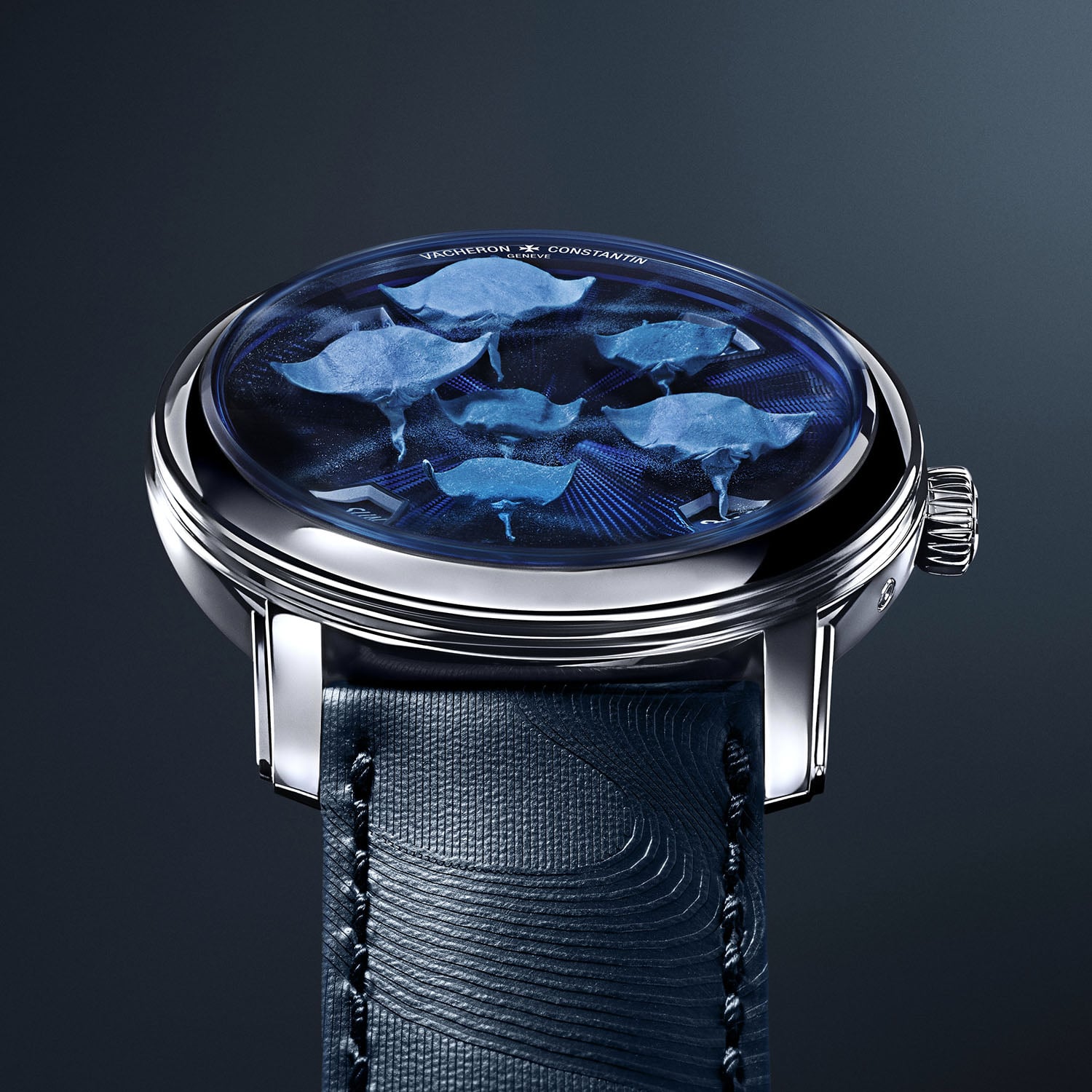 The Oraygami dial seen at an angle, six blue paper manta rays folded in origami across a blue guilloché ground in a white gold case, the four time apertures set into the chapter