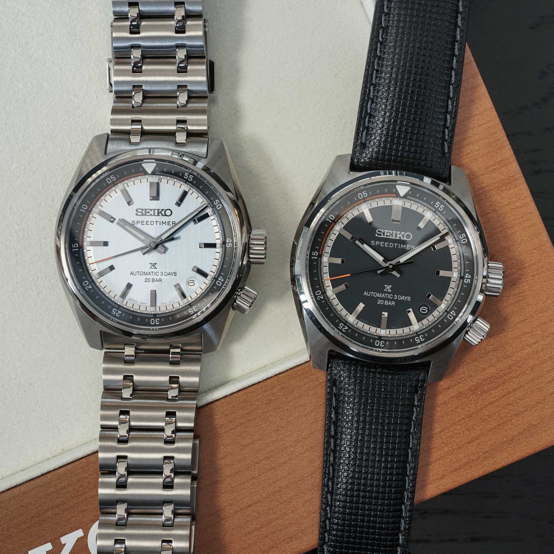 The two Seiko Speedtimer time-and-date models side by side, the light HBC003 on a steel bracelet and the dark-dial HBC004 on a black leather strap, both with inner rotating bezels and orange flange accents