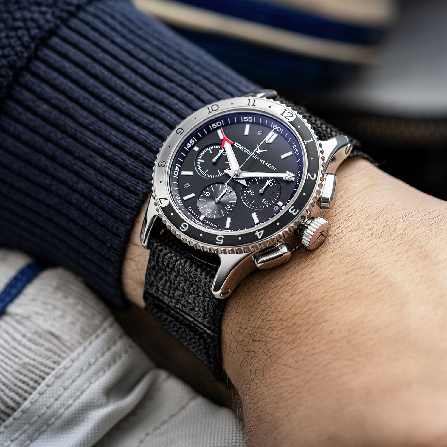 The steel SC02C chronograph worn on a wrist over a navy sweater on a black woven fabric strap, its black dial carrying three subdials and a red-tipped hand